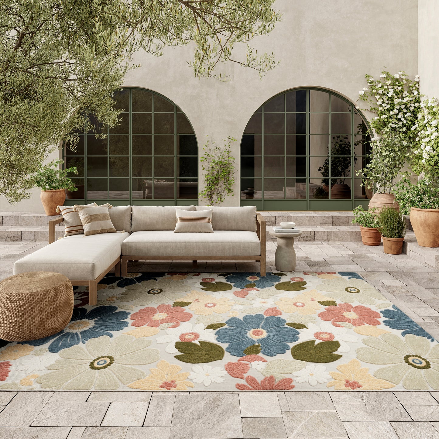 Outdoor patio lounge with beige sectional sofa, vibrant floral rug, terracotta pots, & arched green-framed glass doors.