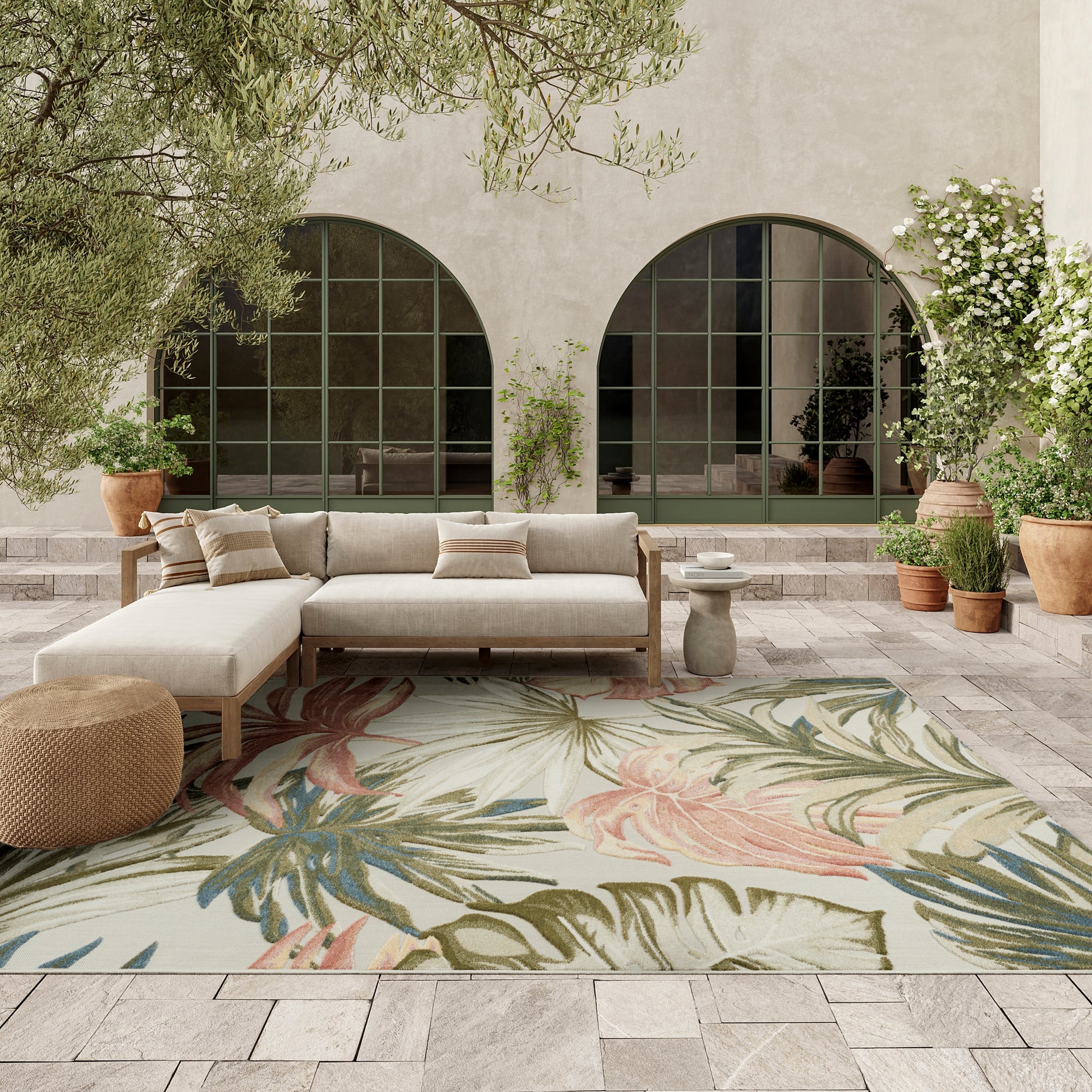 Neutral outdoor lounge with beige sectional on wood frame, tropical leaf rug, potted plants, arched green-framed windows