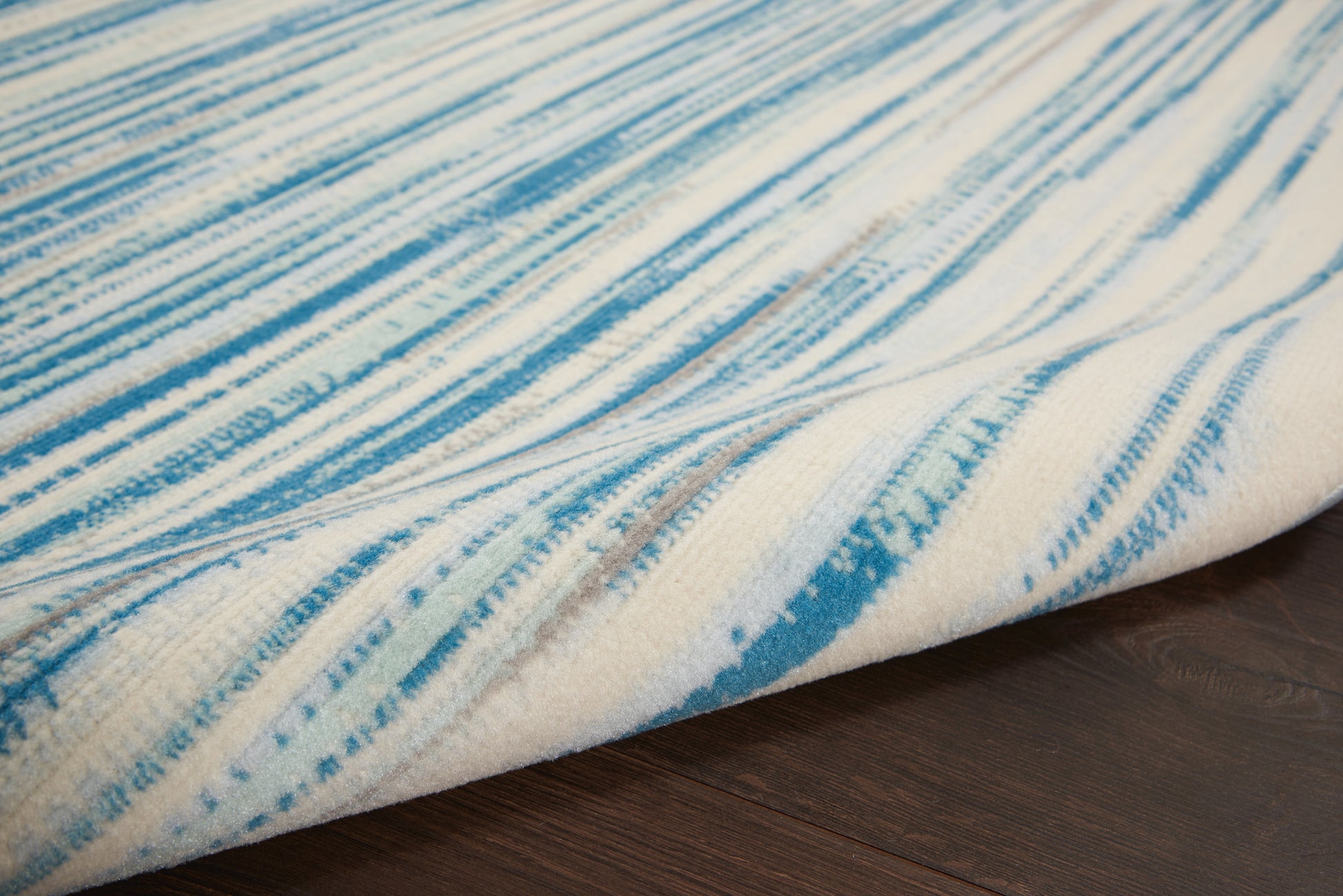 Blue brushstroke striped rug in white/cream tones, soft textured weave; rectangular with folded edge on dark wood floor.