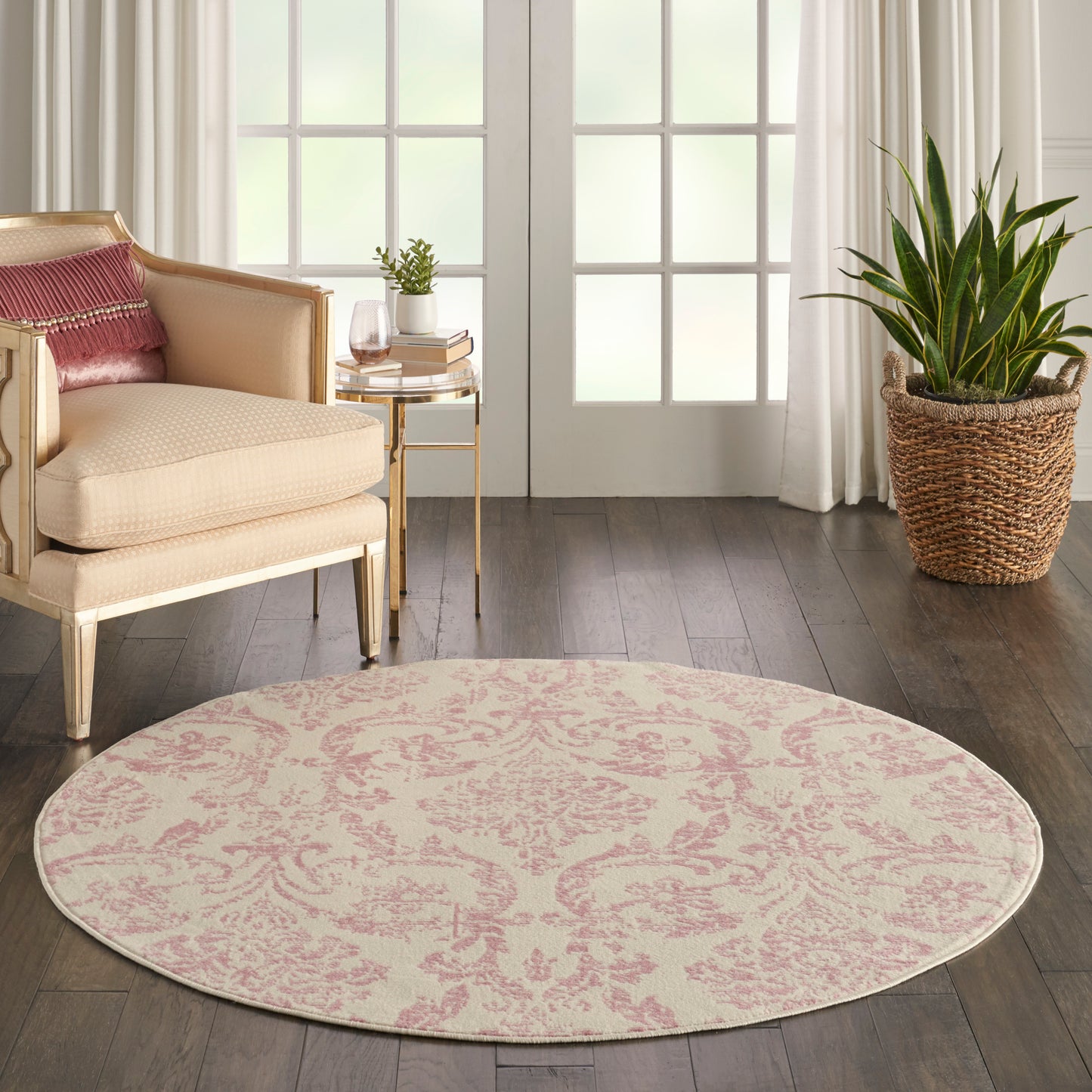 Round pink damask rug on dark wood floor beside beige armchair, gold side table, woven plant basket, and white curtains.