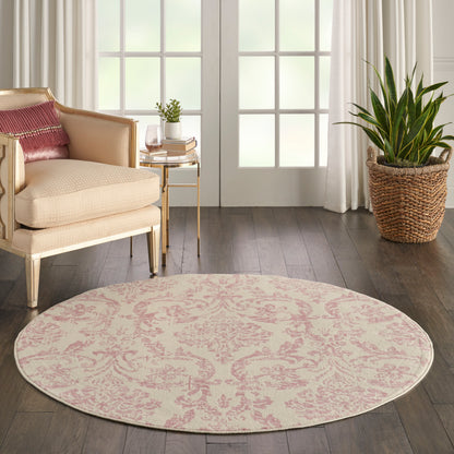Round pink damask rug on dark wood floor beside beige armchair, gold side table, woven plant basket, and white curtains.