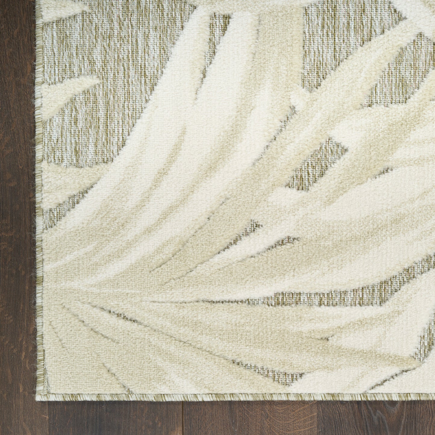 Cream palm-frond pattern area rug with textured beige motif and fringed border on dark wood floor.