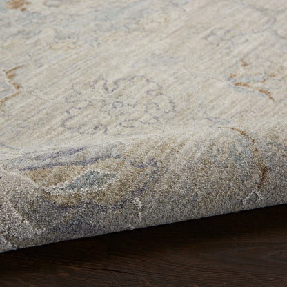 Beige/gray patterned area rug with subtle floral medallion, medium pile, edges curling slightly, over dark hardwood floor.