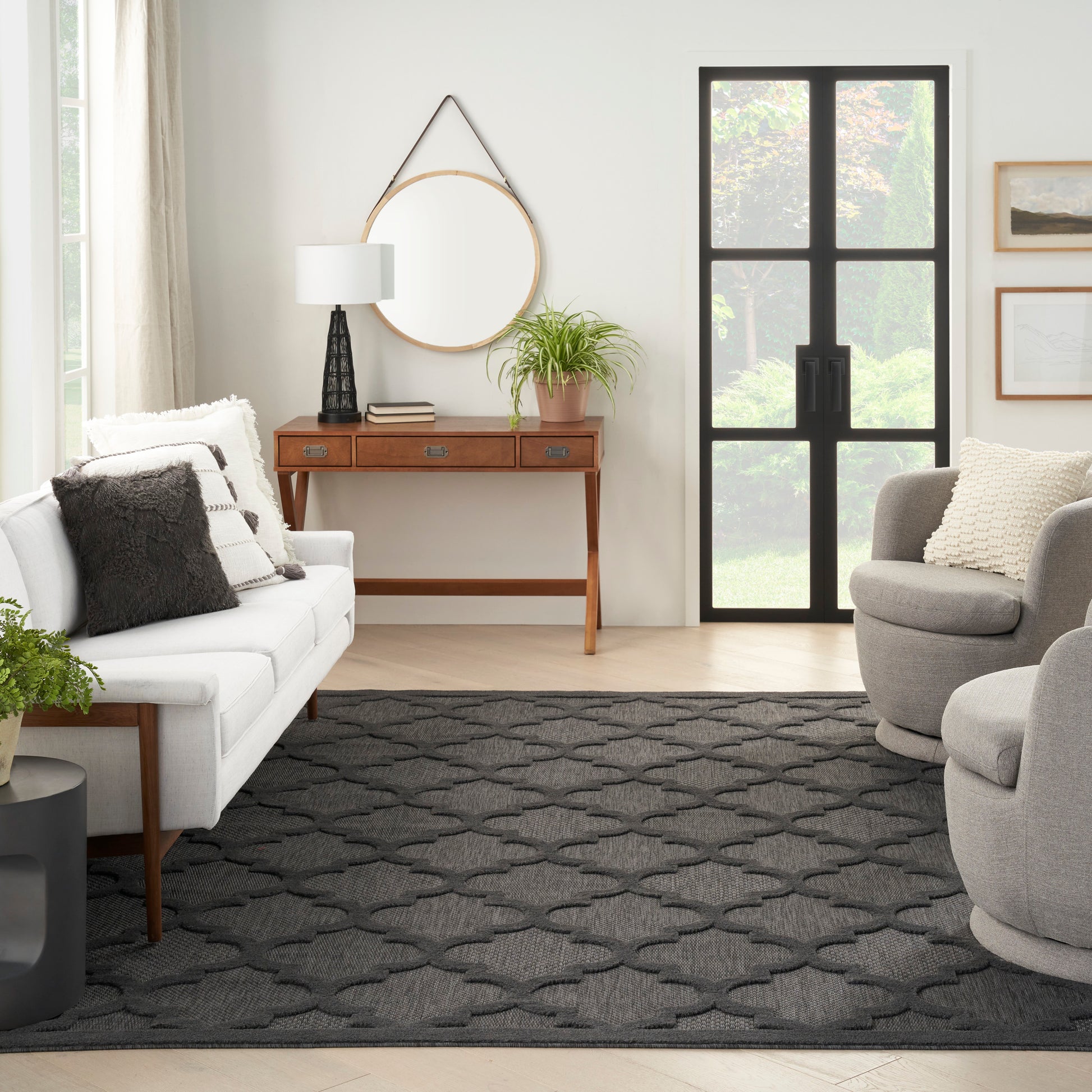 Bright living room with white sofa, gray accent pillows, round mirror above a wooden console, lamp, plant, and dark geometric rug.