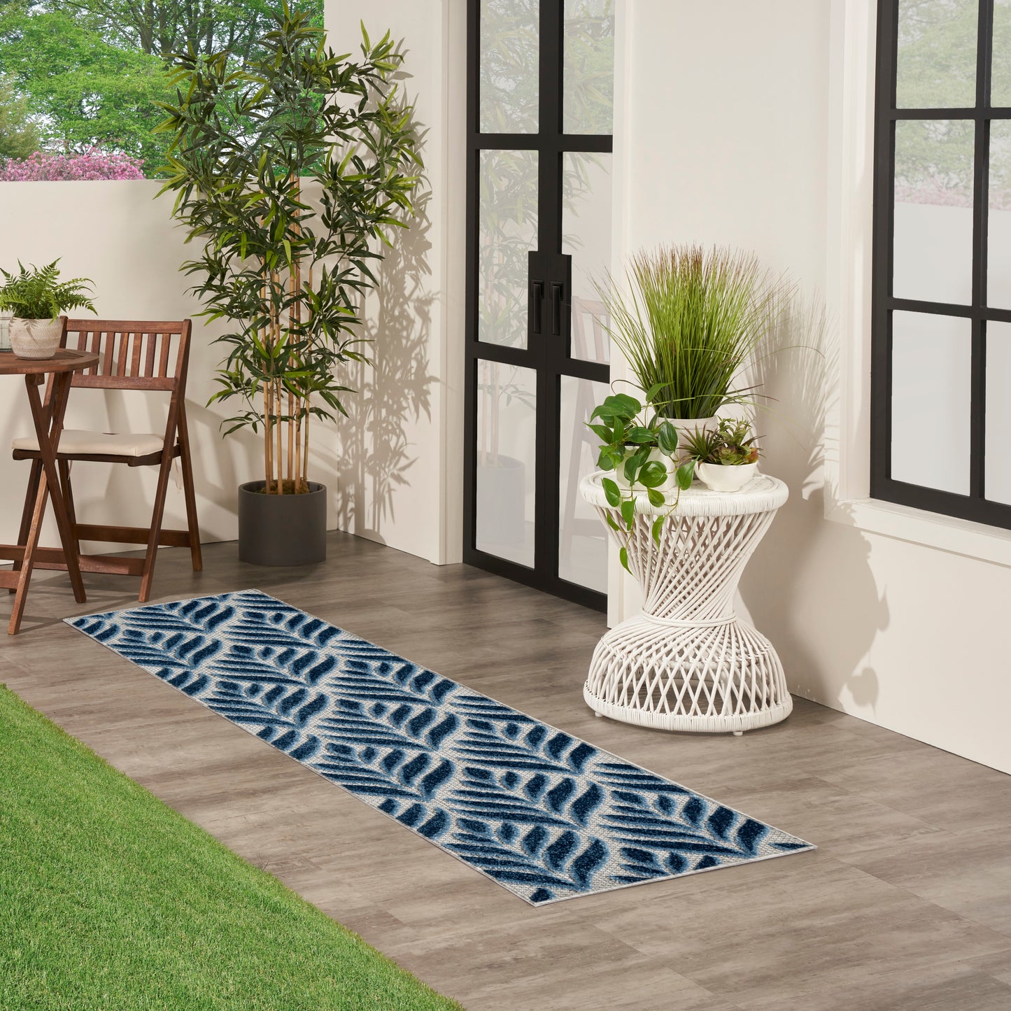 Blue leaf-pattern runner rug on wood floor beside white wicker plant stand and glass doors with potted greenery.