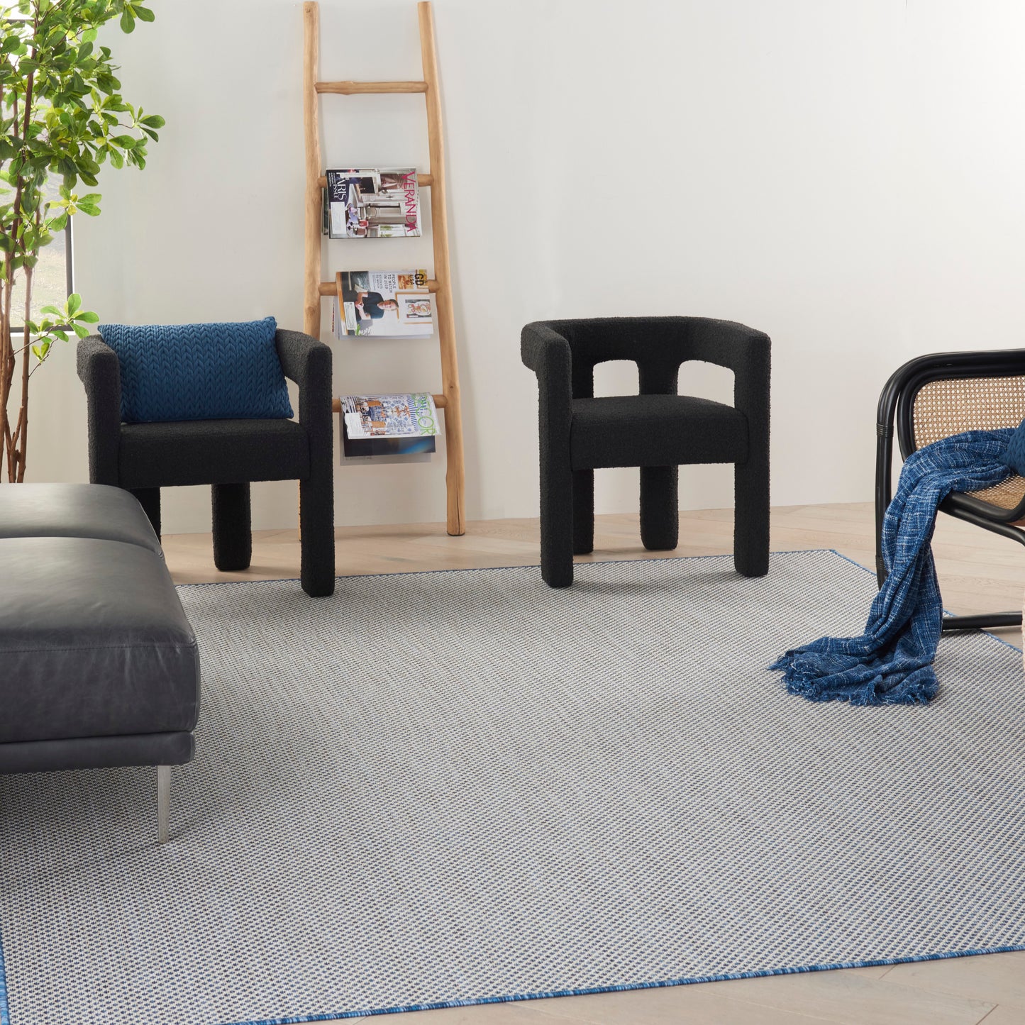Modern living room with black fabric sculptural lounge chairs, blue cushion, wooden ladder shelf, green plant, and gray area rug.