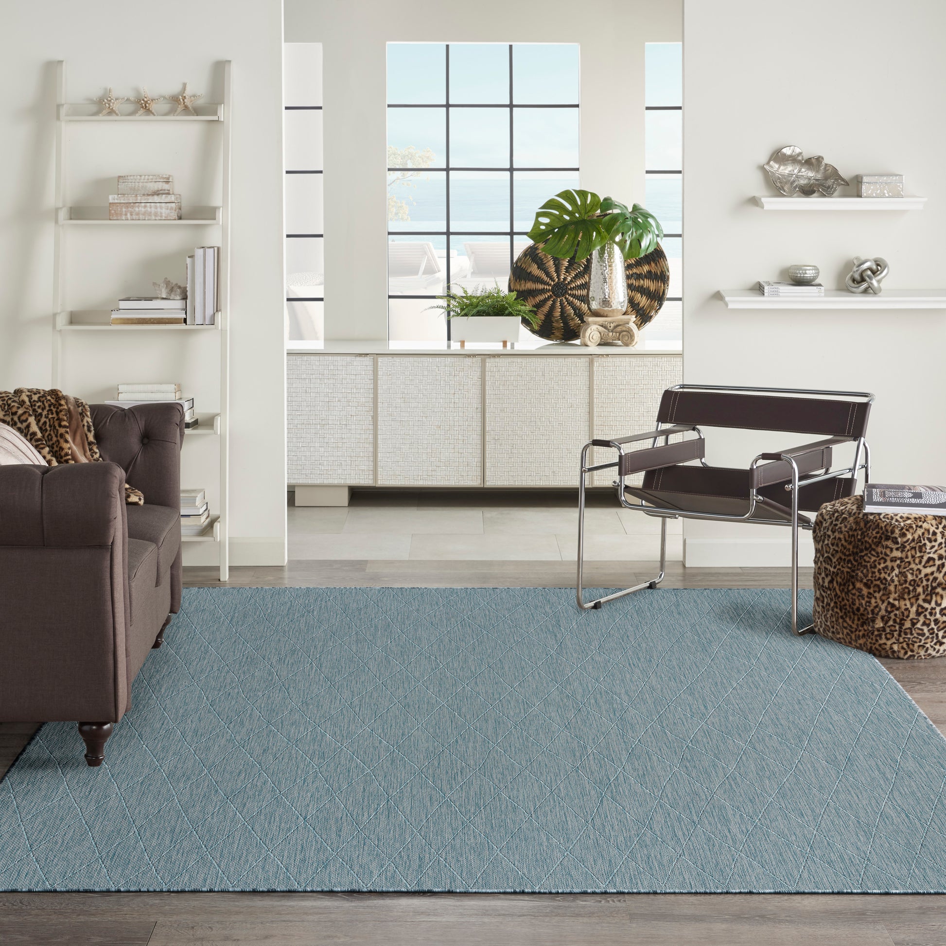 Modern living room with a light blue diamond rug, brown leather sofa, metal lounge chair, leopard ottoman, white shelves.