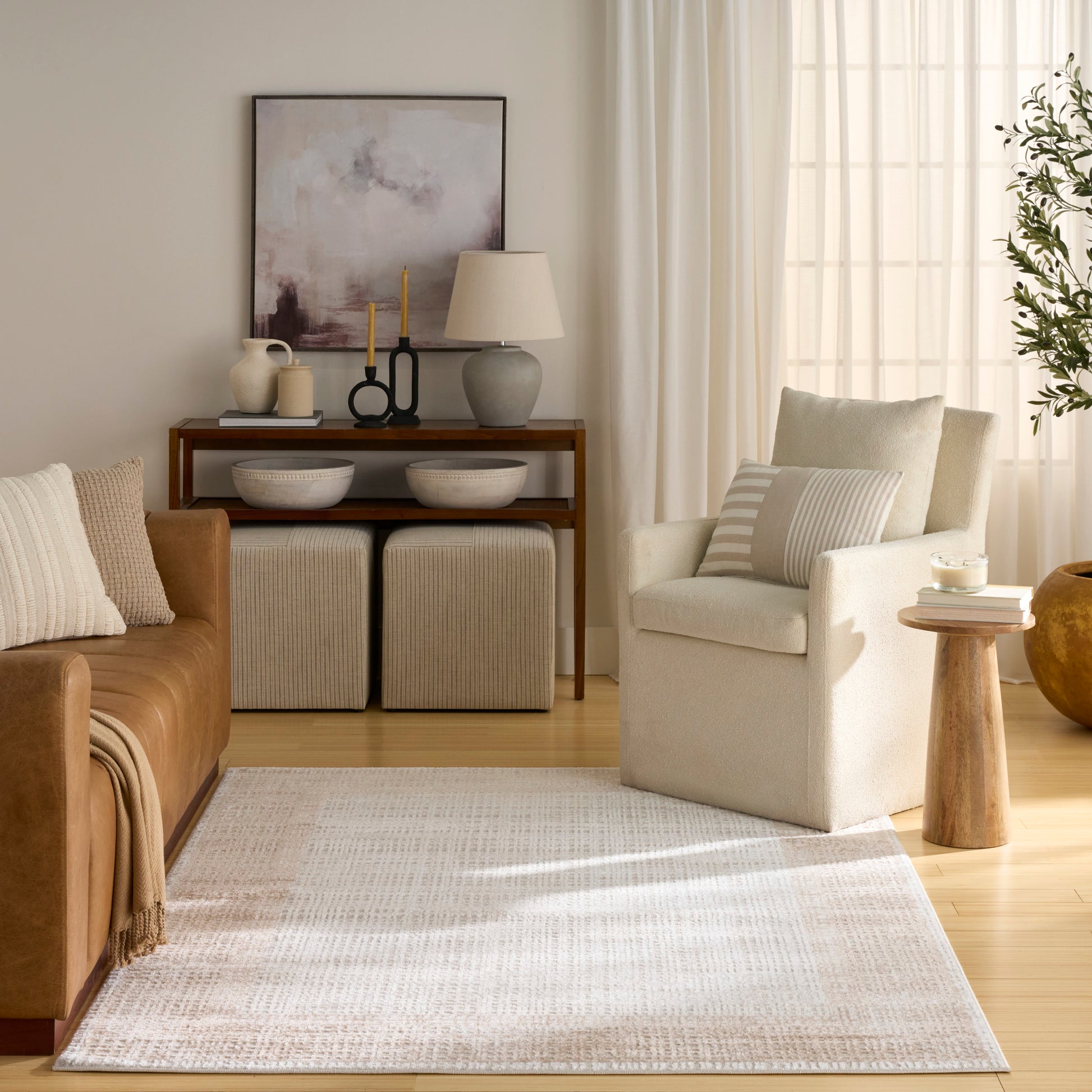 Neutral living room with cream textured rug, tan leather sofa, beige armchair, wooden console and woven ottomans.