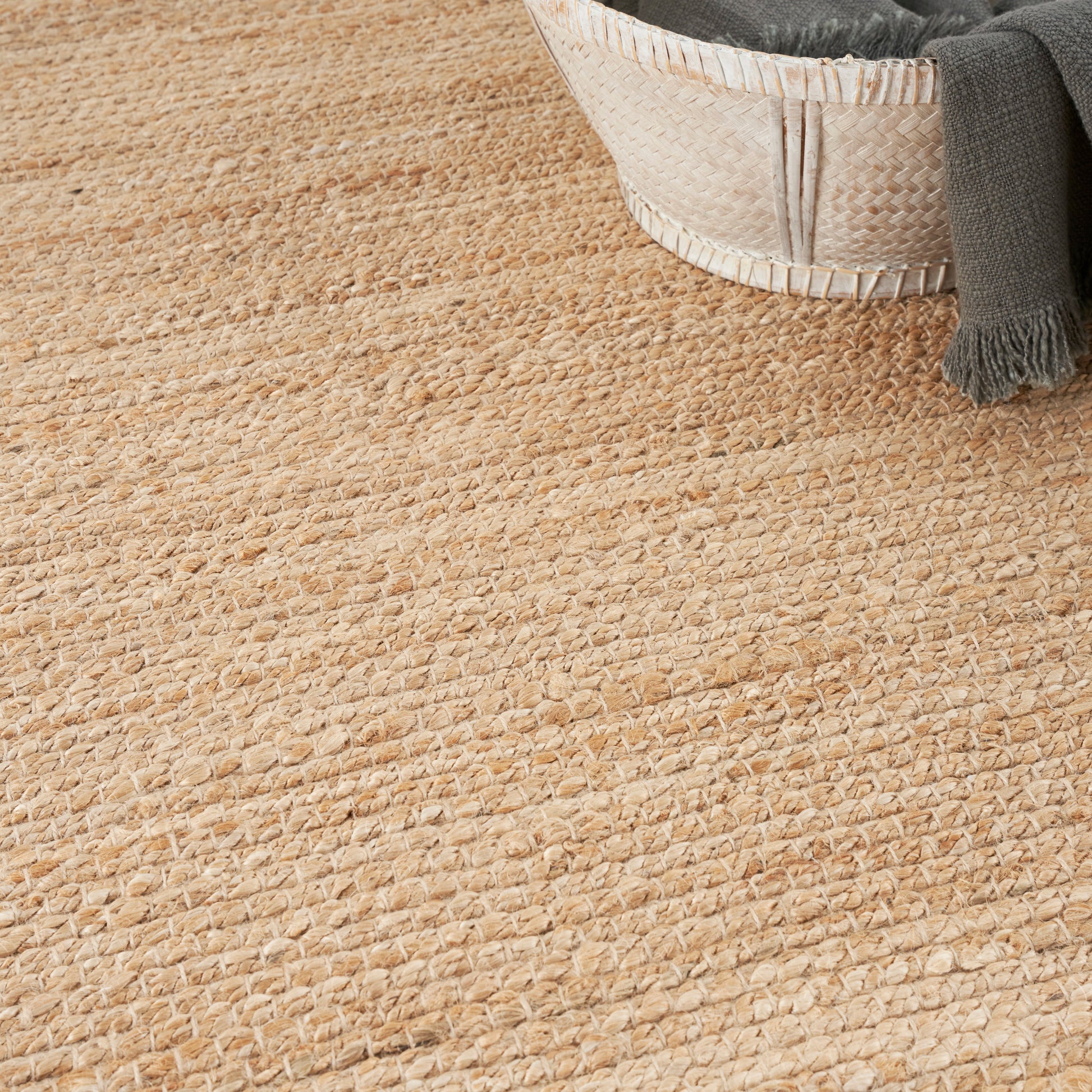 Rectangular beige natural jute rug with textured loop weave; gray throw draped over a woven basket.