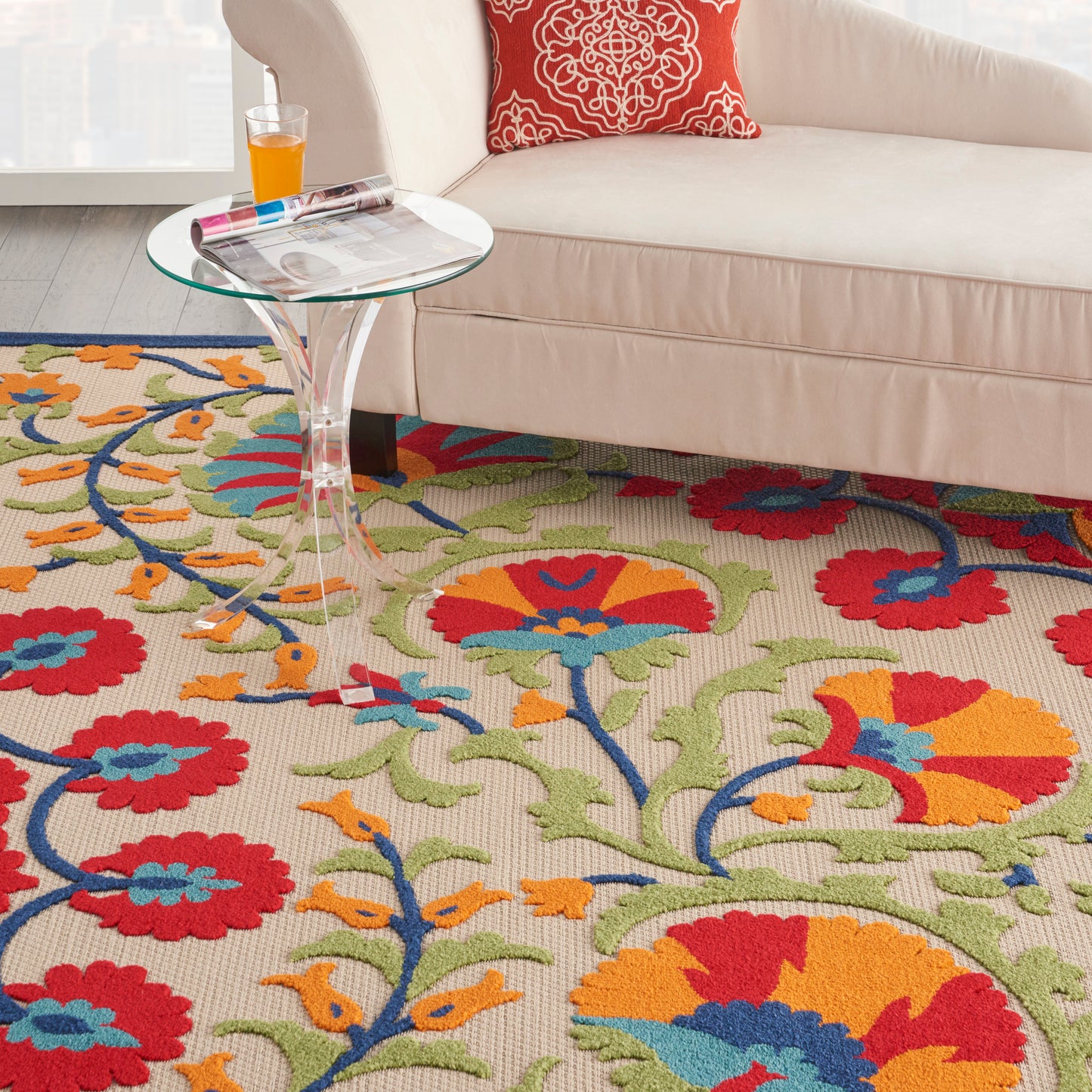 Bright floral area rug in red, orange, blue, green under a glass round side table beside a beige sofa.