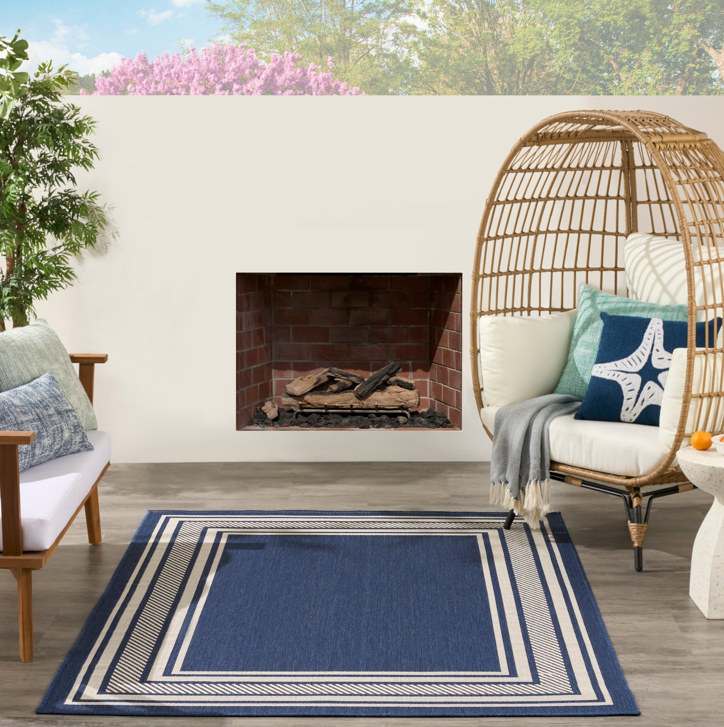 Blue outdoor rug with cream borders on patio, wicker dome chair with cream cushions and starfish pillow, brick fireplace.