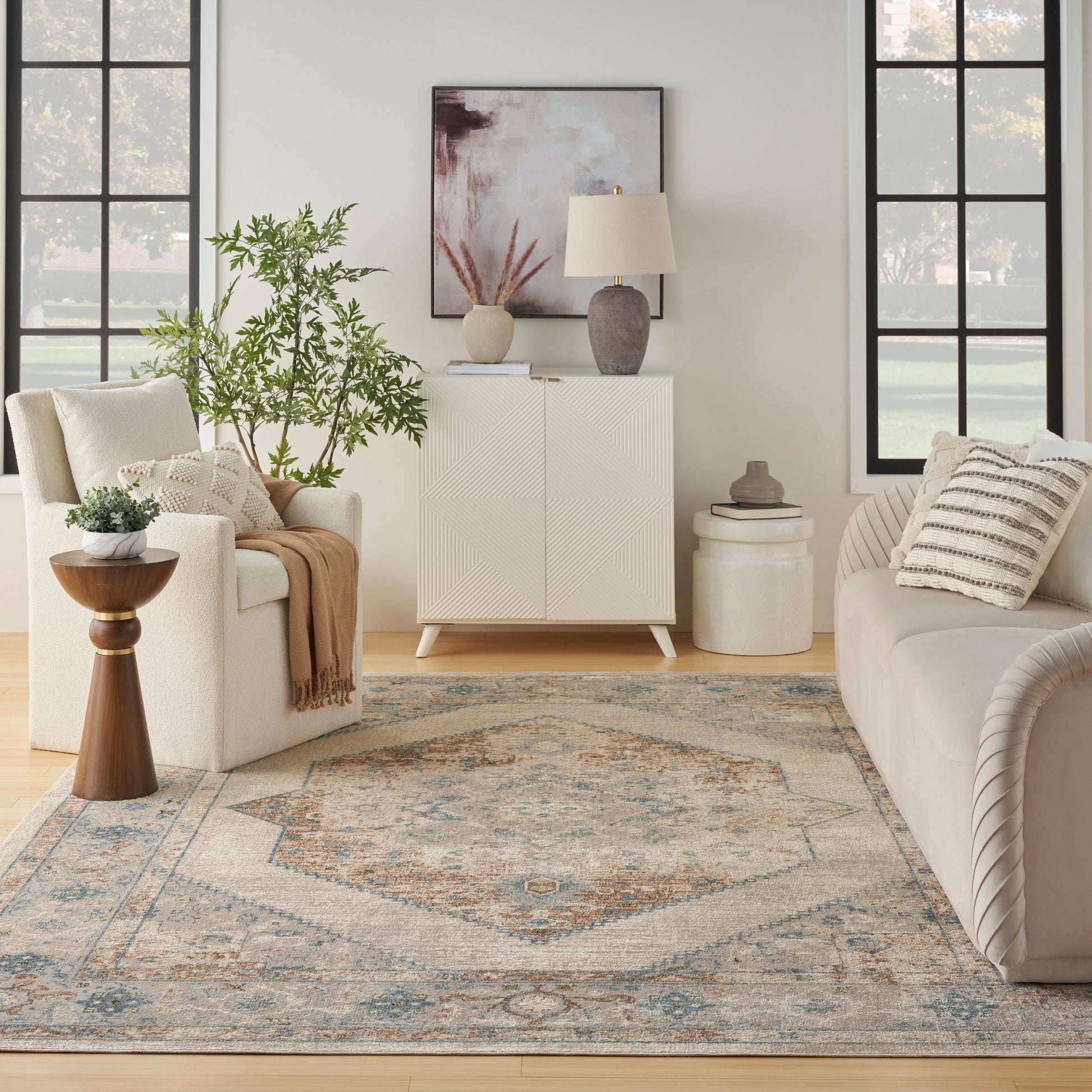 Bright living room with cream sofas, white cabinet, abstract art, ceramic lamp, plant, and blue-beige geometric rug.