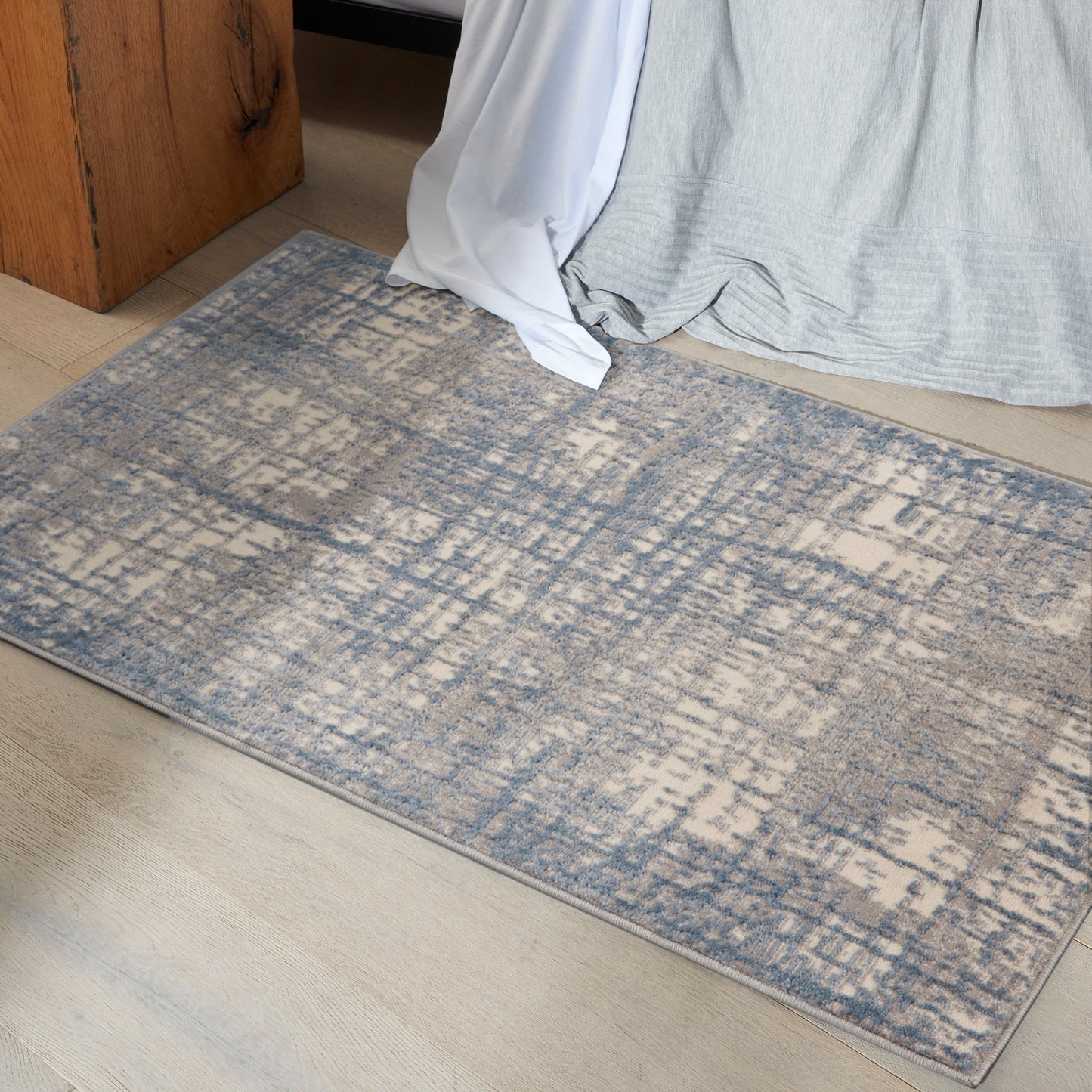 Rectangular low-pile rug with blue and beige distressed abstract pattern on light wood floor beside a bed.