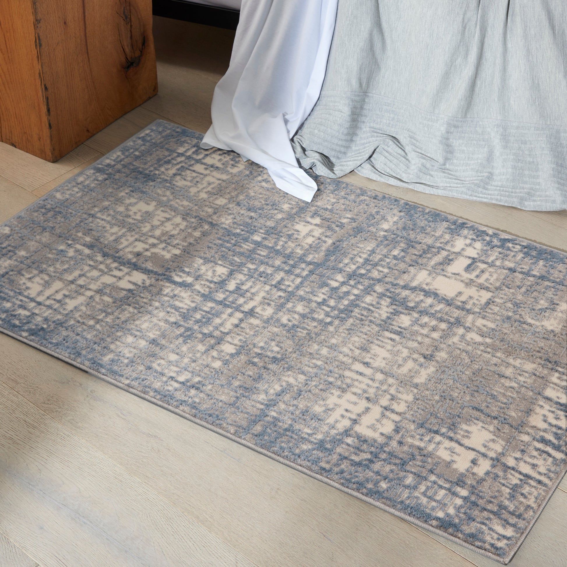 Rectangular low-pile rug with blue and beige distressed abstract pattern on light wood floor beside a bed.