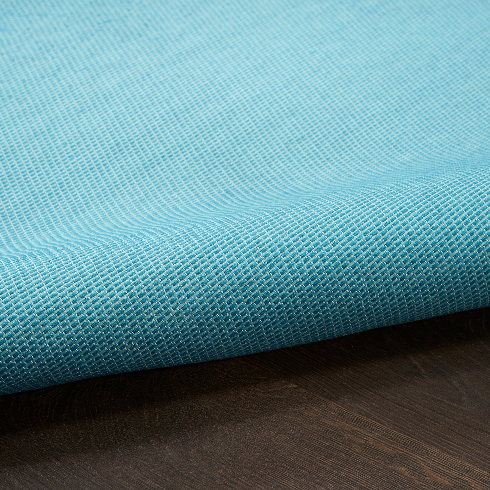 Turquoise woven rug with a white grid pattern, rolled on a dark wood surface.