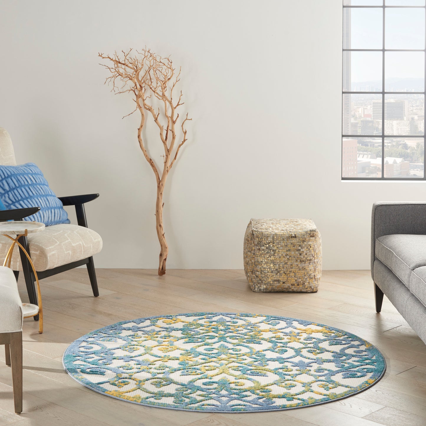 Round blue and teal patterned rug with yellow accents in a bright living room; driftwood tree, woven pouf, gray sofa.