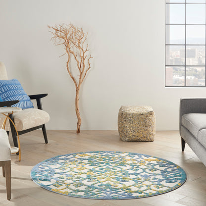 Round blue and teal patterned rug with yellow accents in a bright living room; driftwood tree, woven pouf, gray sofa.