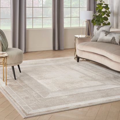 Large light gray textured area rug with a subtle border, set in a modern beige living room with sofa and plant.