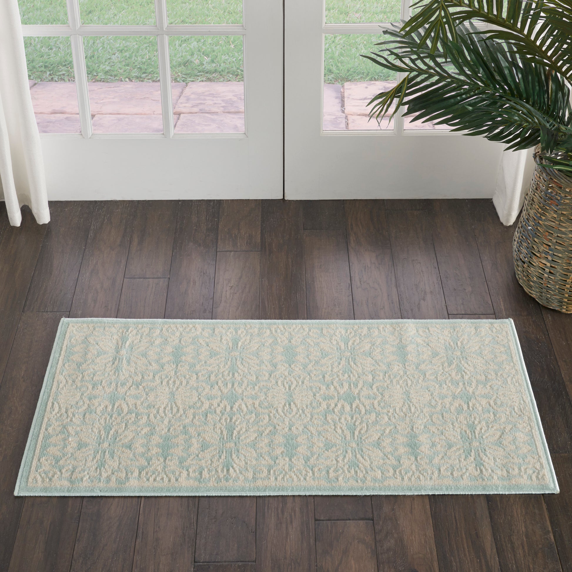 Rectangular light cream rug with pale blue damask pattern and pale blue border on dark hardwood floor near glass doors.