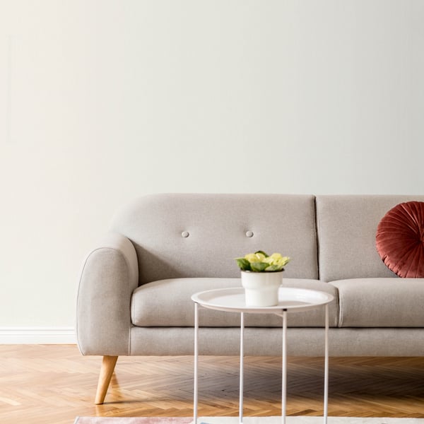 Beige fabric sofa with wooden legs, round white side table with a small plant, and a red cushion on a light wood floor.