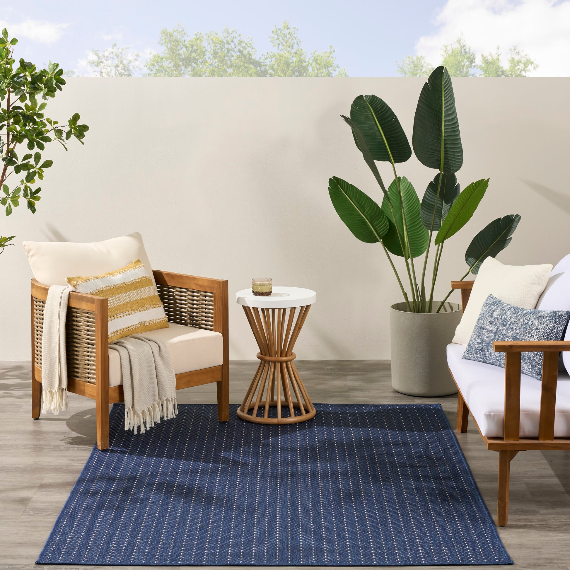 Outdoor wooden-framed chairs with beige cushions, navy striped rug, round wicker side table, and large potted tropical plants.