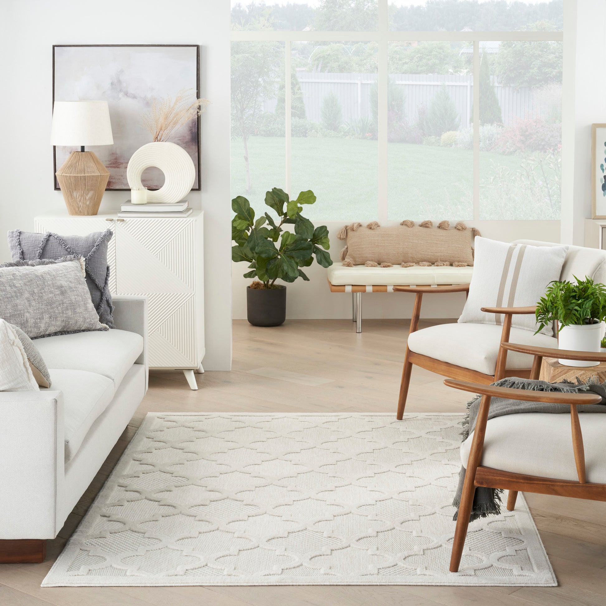 Bright living room with white sofa, light wood furniture, patterned beige rug, potted plants, and a wooden-framed chair.