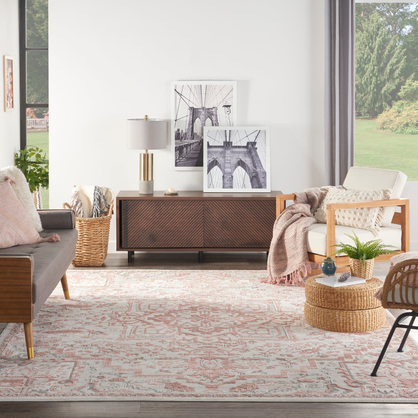 Mid-century living room: gray sofa, wood sideboard, black-and-white prints, woven ottoman, pink rug, throws, large windows.