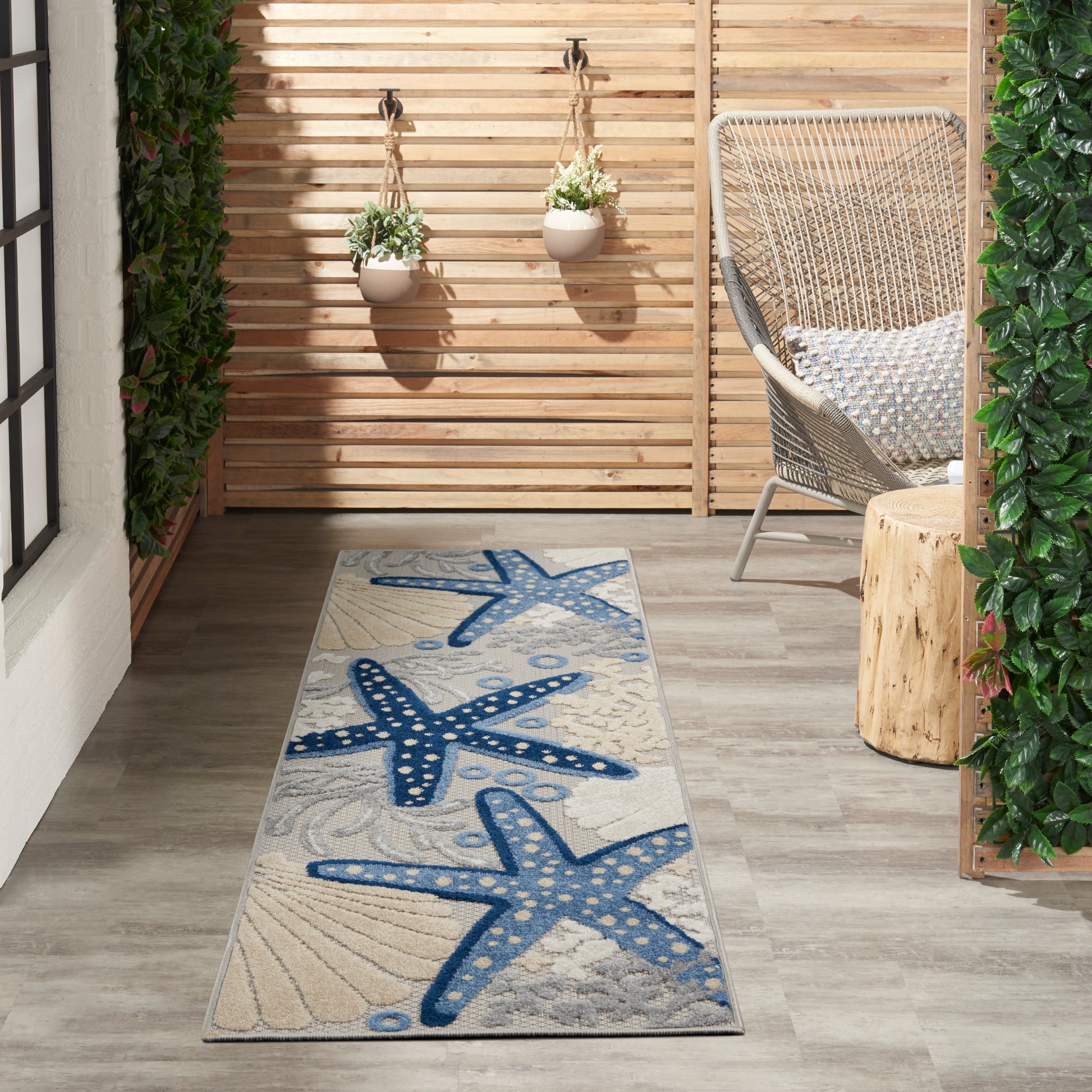 Coastal outdoor runner rug featuring a blue starfish pattern on beige, set on a gray wooden deck with a wicker chair and planters