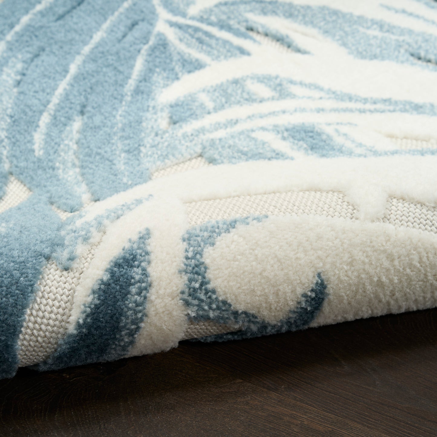 Blue & white plush shag rug with abstract pattern, rolled edge showing underside, on dark wood floor.