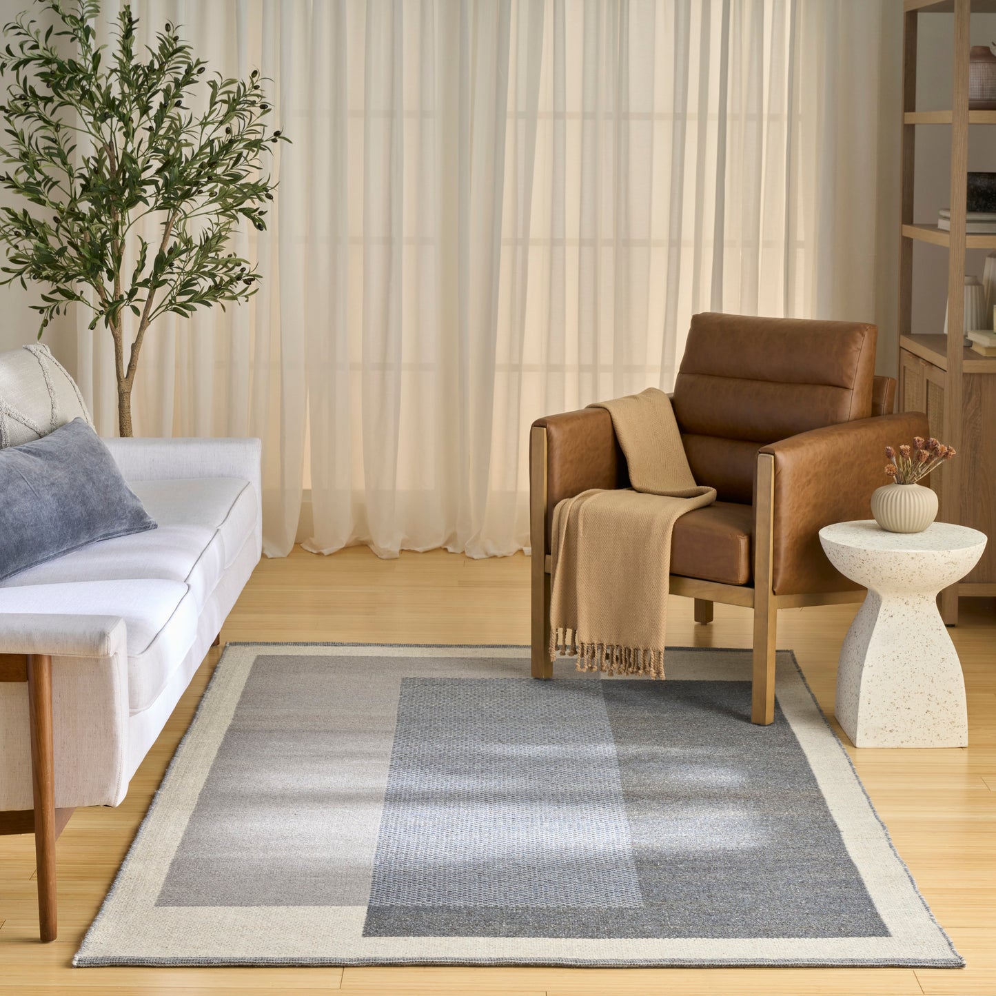 Cozy living room with gray patchwork rug, white sofa, brown leather armchair, beige throw, plant, and light wood floor.