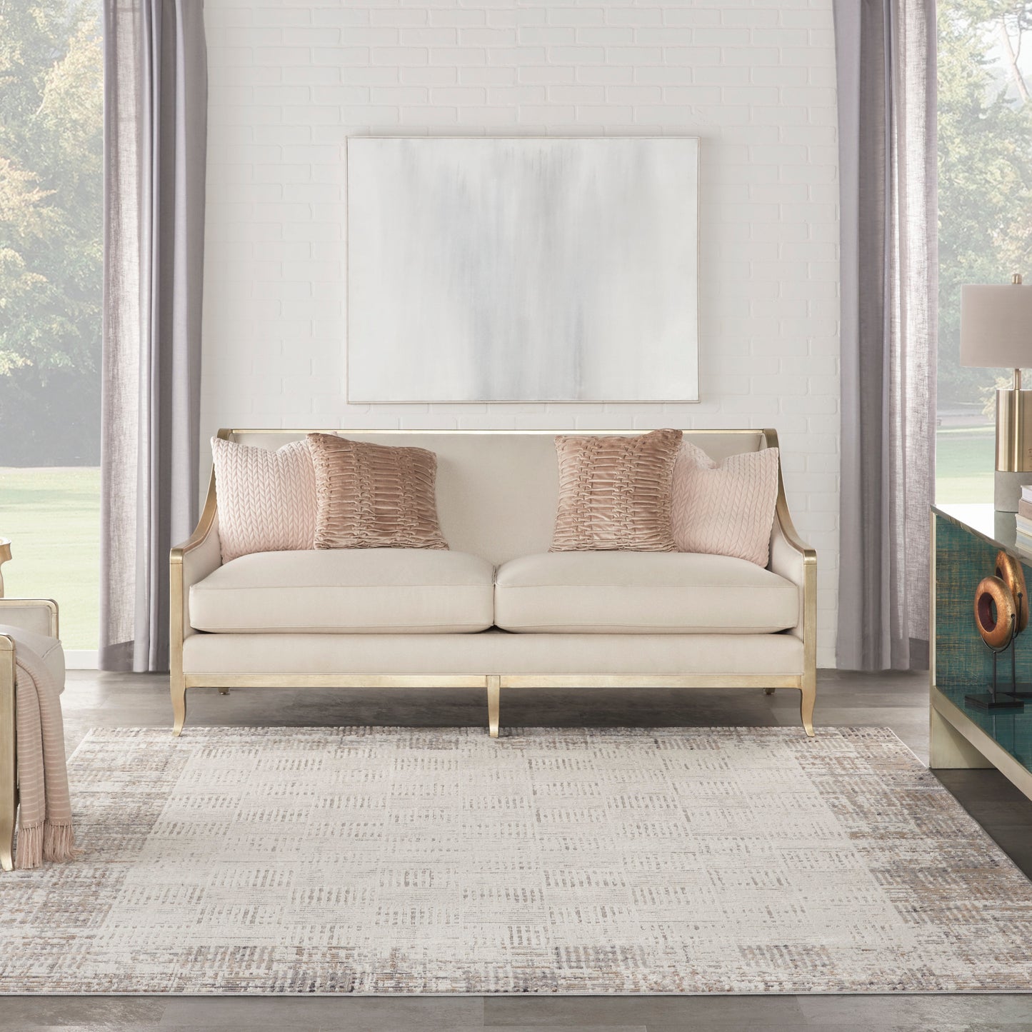 Cream sofa with gold frame & pink textured pillows in bright living room with abstract art, white brick wall & light gray rug.