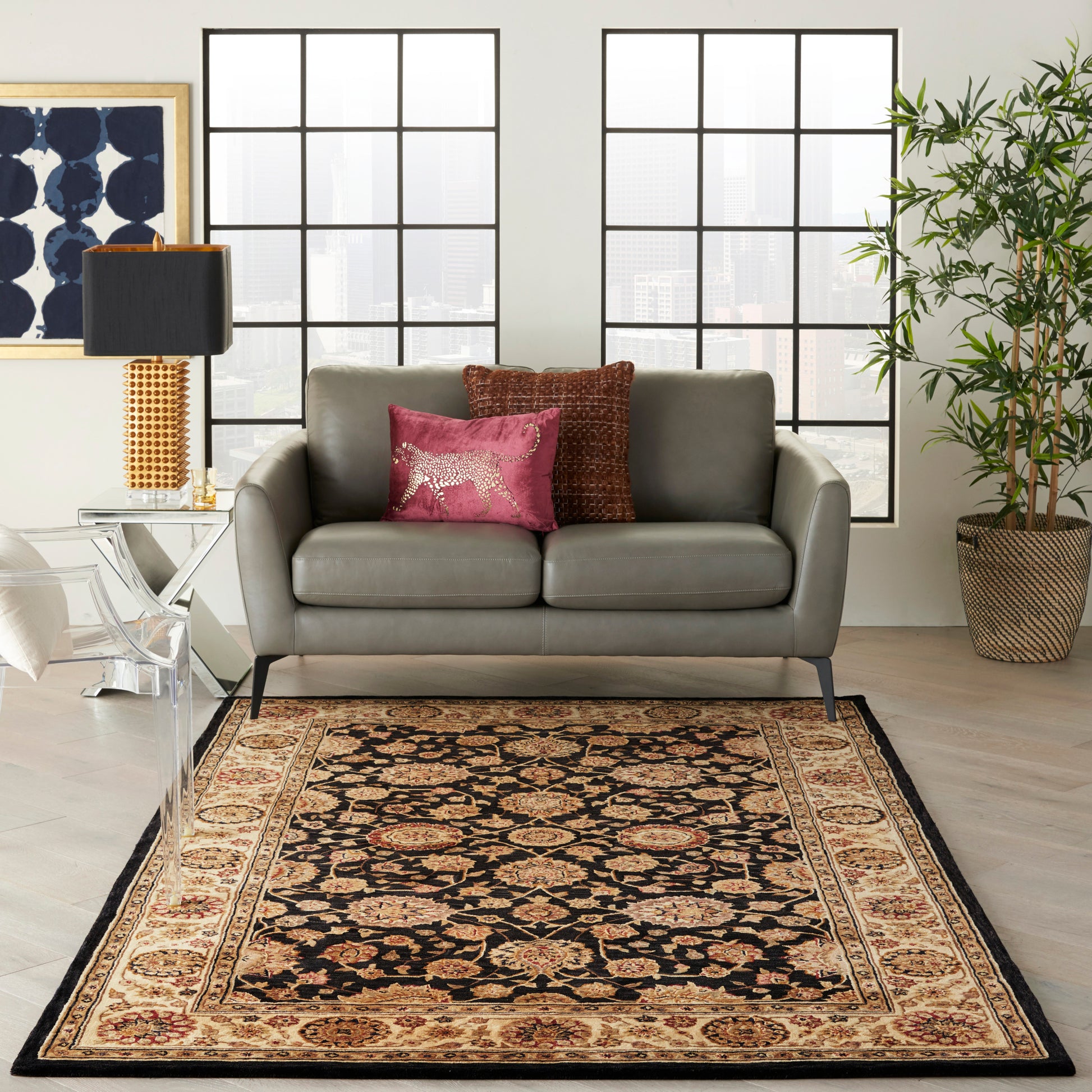 Gray leather sofa on a black-beige oriental rug; pink and copper cushions, acrylic chair, glass side table, tall lamp, potted plant.