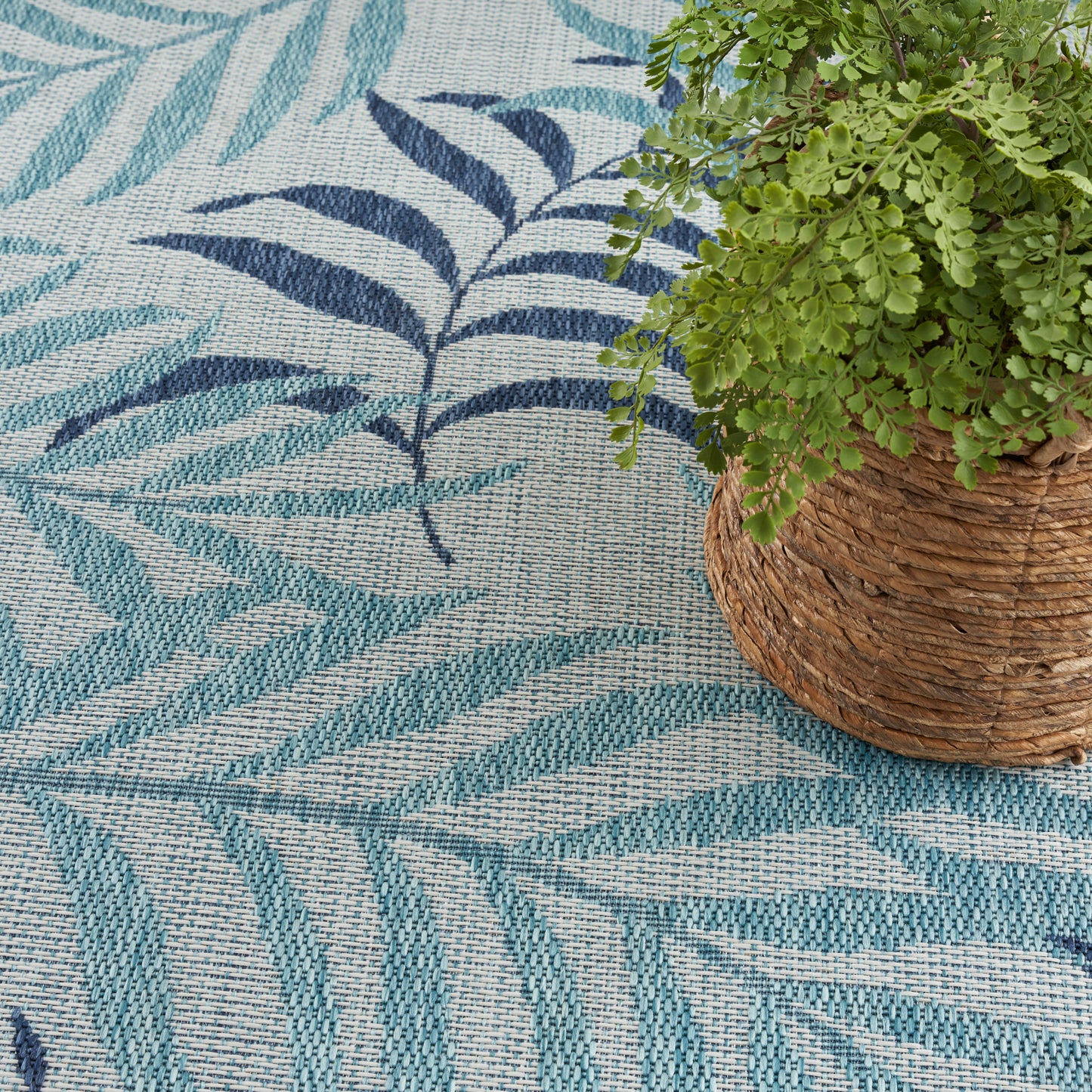 Teal & navy leaf-patterned woven rug with a tropical vibe, paired with a potted fern in a rustic wicker basket.