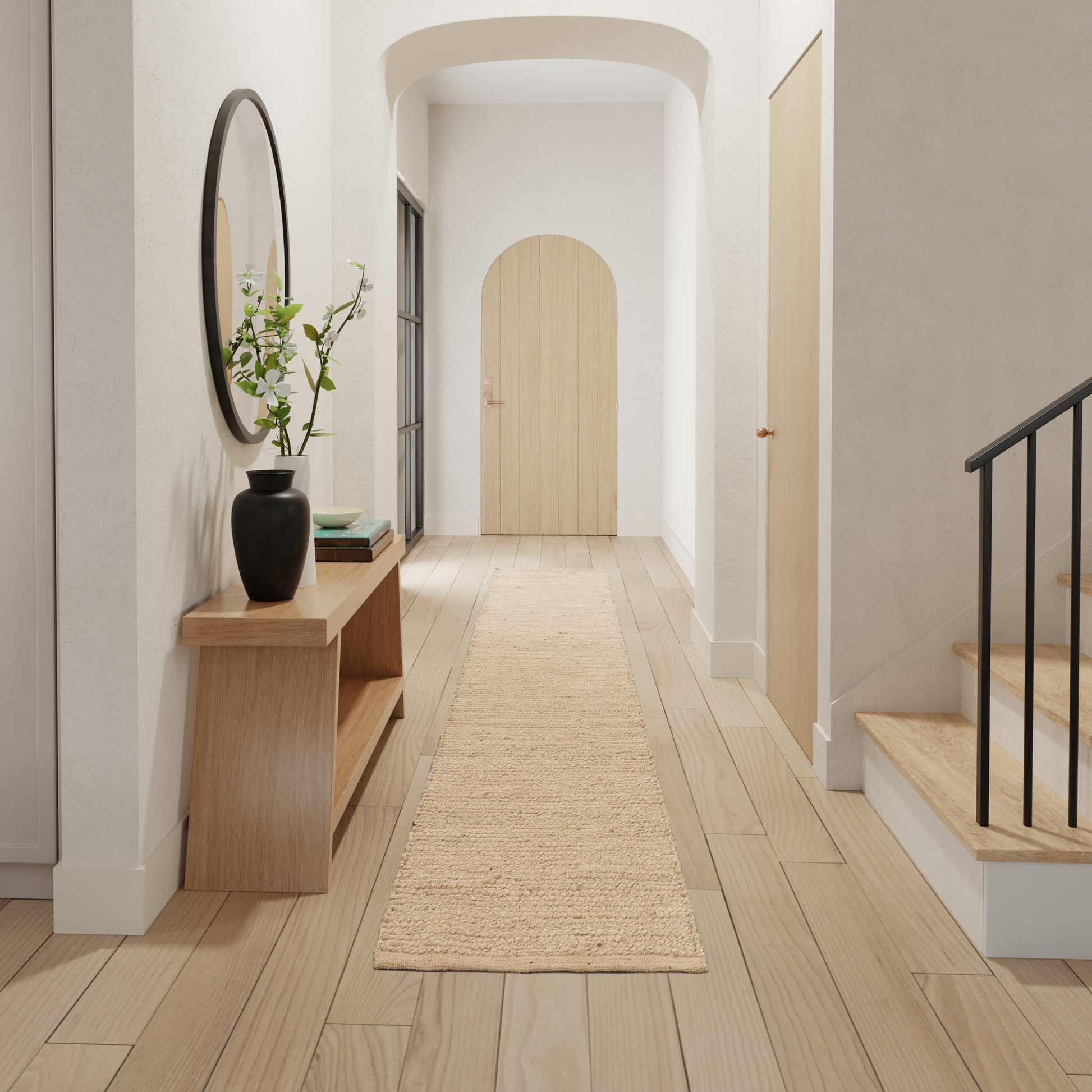 Minimalist light-wood hallway with arched doorway, woven jute runner, console table, black vase, round mirror, and stairs.
