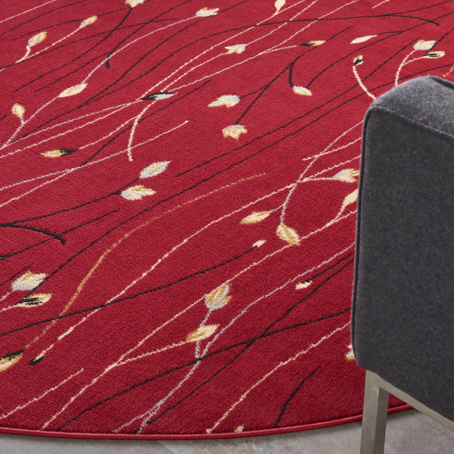 Red circular rug with cream leaf accents and black linear streaks, paired with a gray upholstered chair.