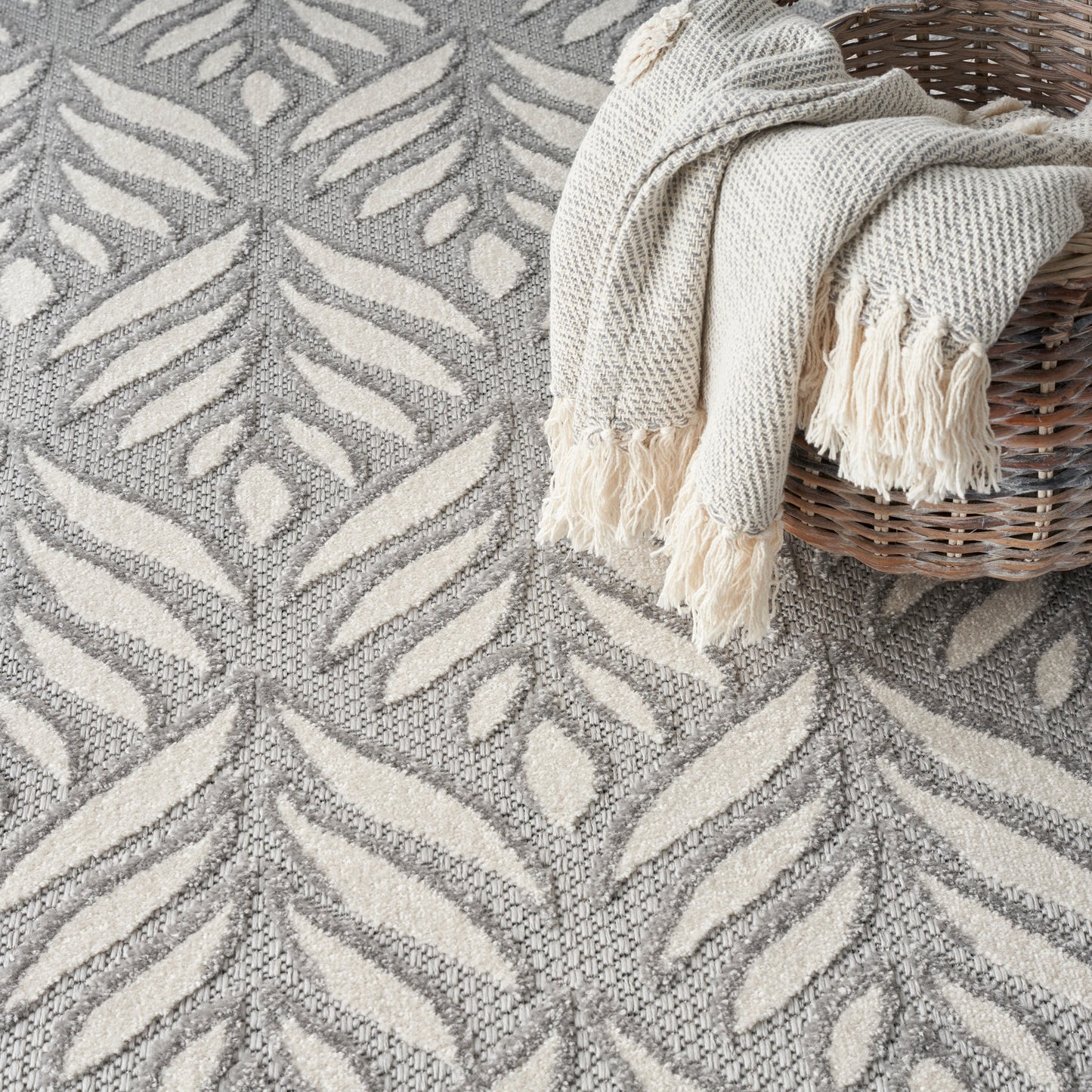 Grey & ivory leaf-patterned woven rug with a cream fringed throw draped over a wicker basket.