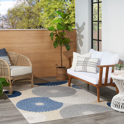 Outdoor patio with wicker rattan chairs and a wooden-framed sofa, white cushions, blue-gray rectangular 5' x 8' rug, potted plant, wicker side table.