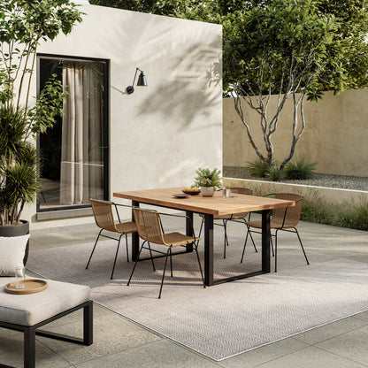 Outdoor dining set with rectangular wooden table and six wicker chairs on a gray patterned rug beside beige stucco wall.
