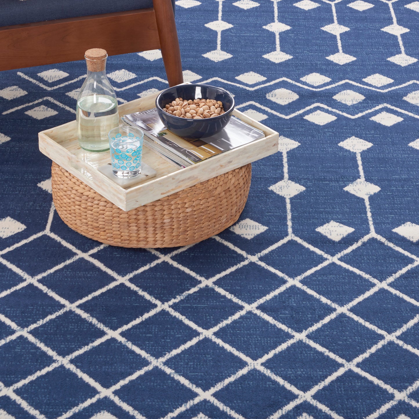 Blue geometric rug featuring a round woven rattan basket on a light wood tray holding a bottle, a blue glass, and a bowl of peanuts.