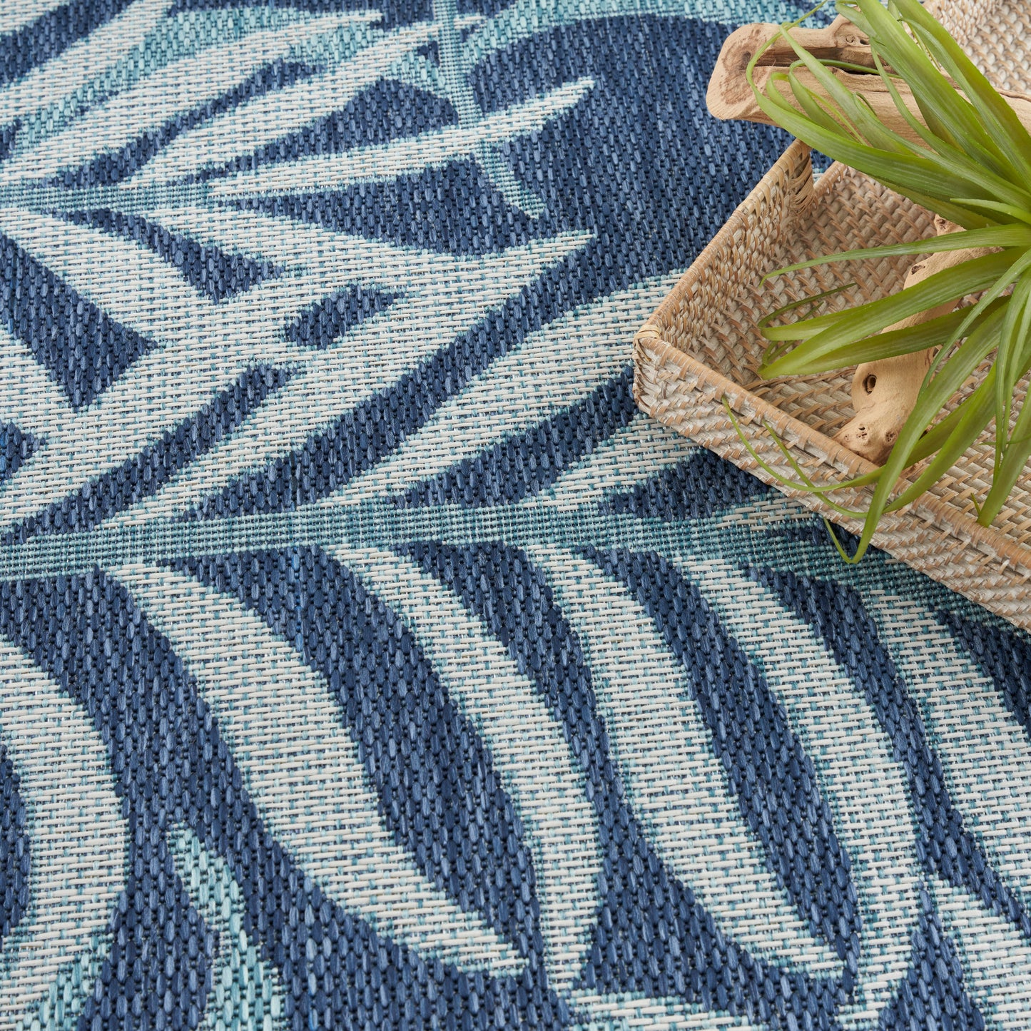 Woven navy and sky-blue rug with bold abstract leaf pattern; natural wicker basket with a green plant on top.