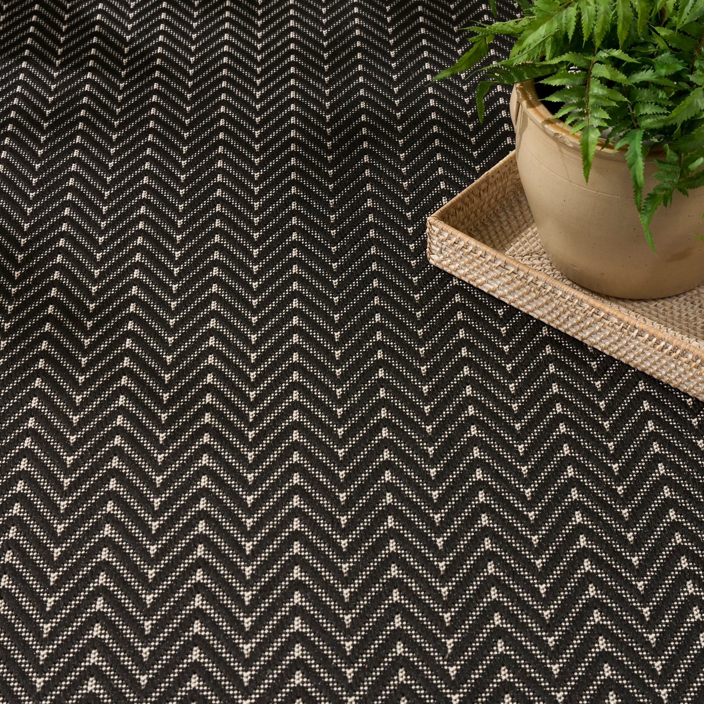 Dark charcoal chevron rug with white specks, beige woven tray, and a green fern in a clay pot.