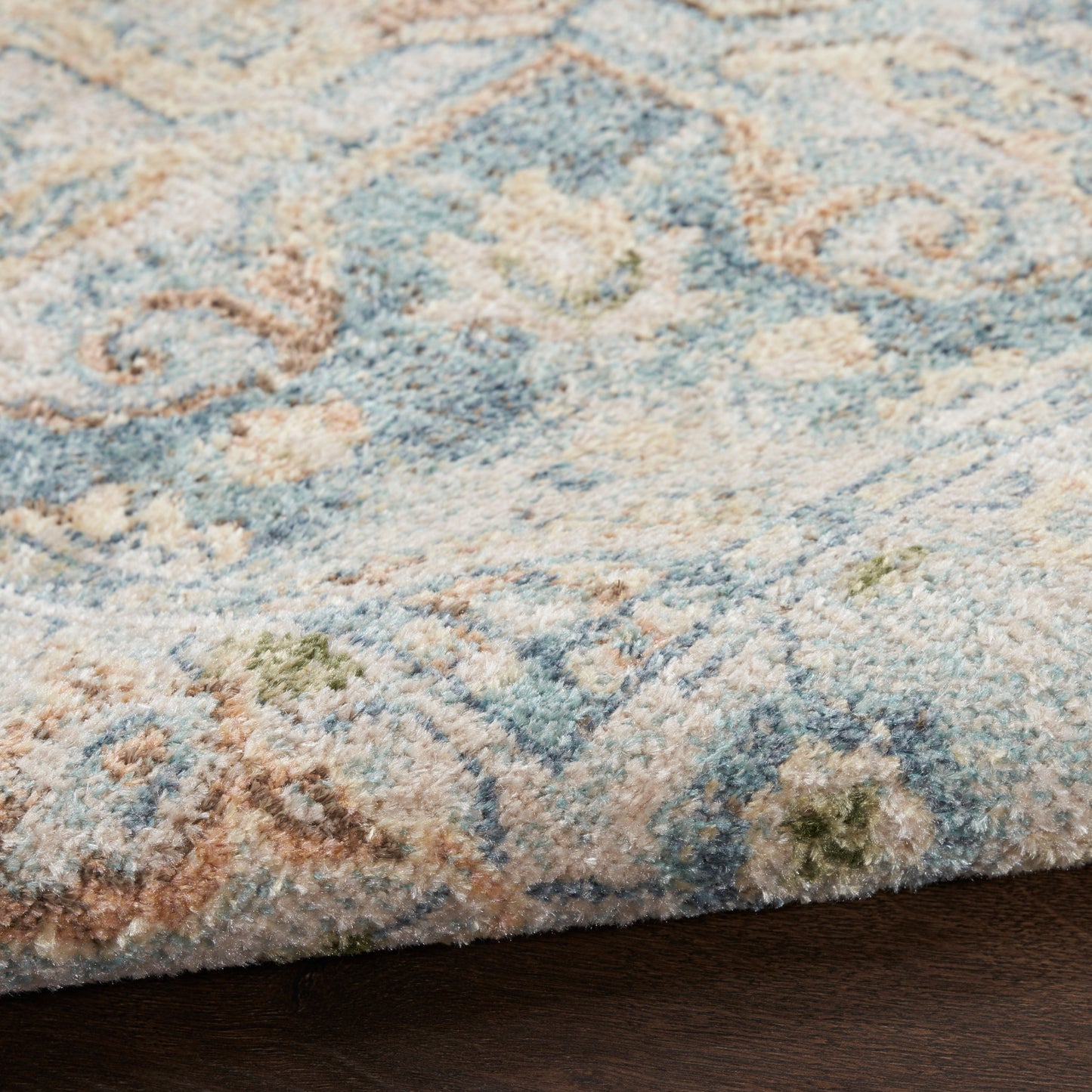 Close-up of pastel blue and cream patterned rug with beige and green accents on dark wood floor.