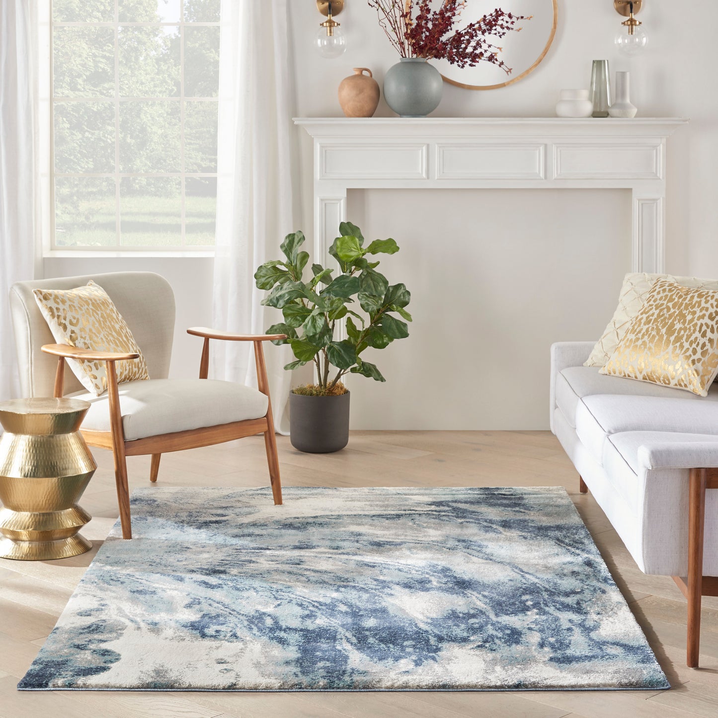 Cozy living room featuring a blue abstract area rug, white sofa, wooden armchair, brass side table, potted plant, and white mantel decor.