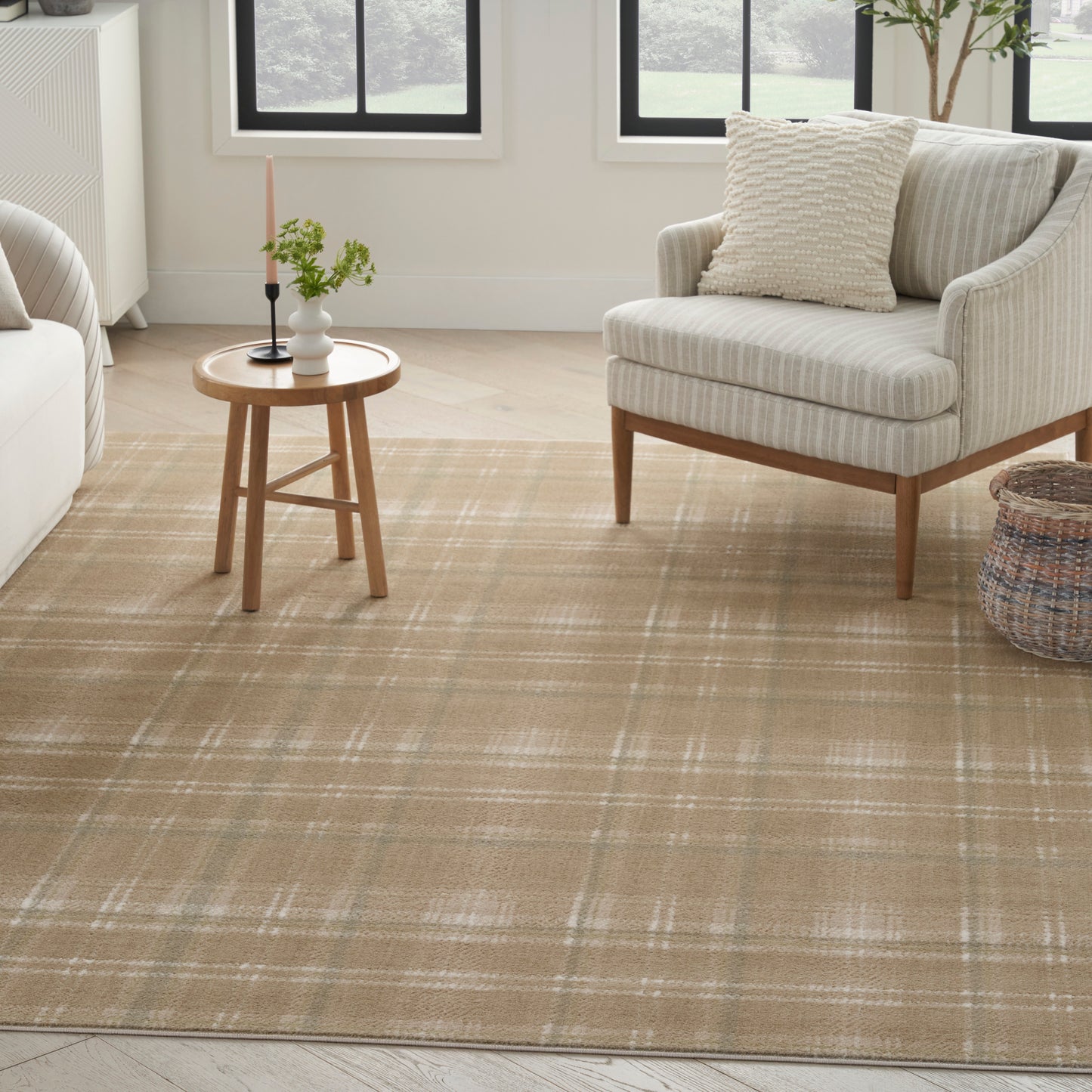 Beige plaid woven area rug in a bright living room with a cream upholstered armchair, round wood side table and basket.