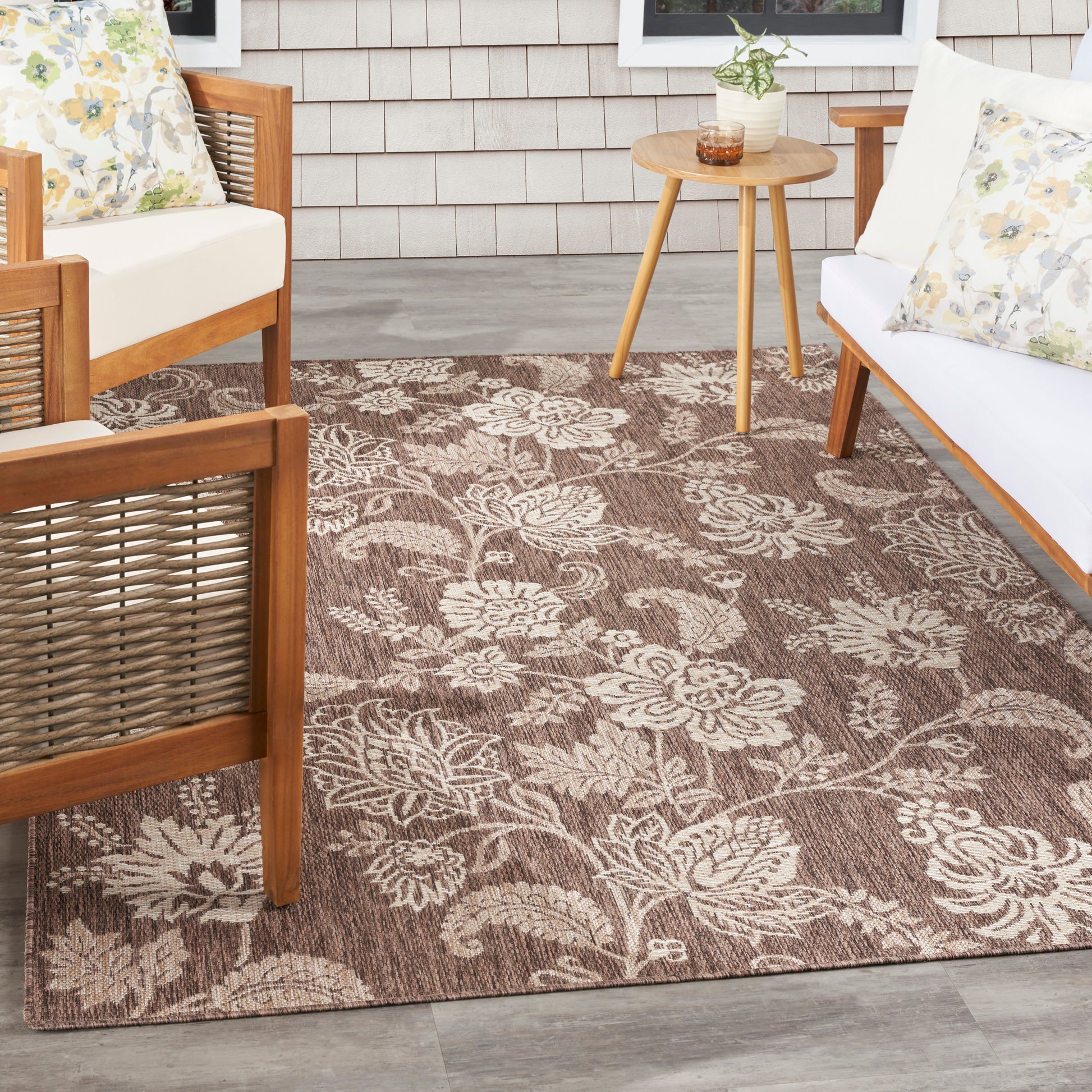 Brown outdoor rug with cream floral pattern on a porch, paired with wooden-frame seating and a round side table.