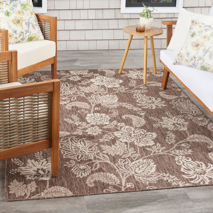 Brown outdoor rug with cream floral pattern on a porch, paired with wooden-frame seating and a round side table.