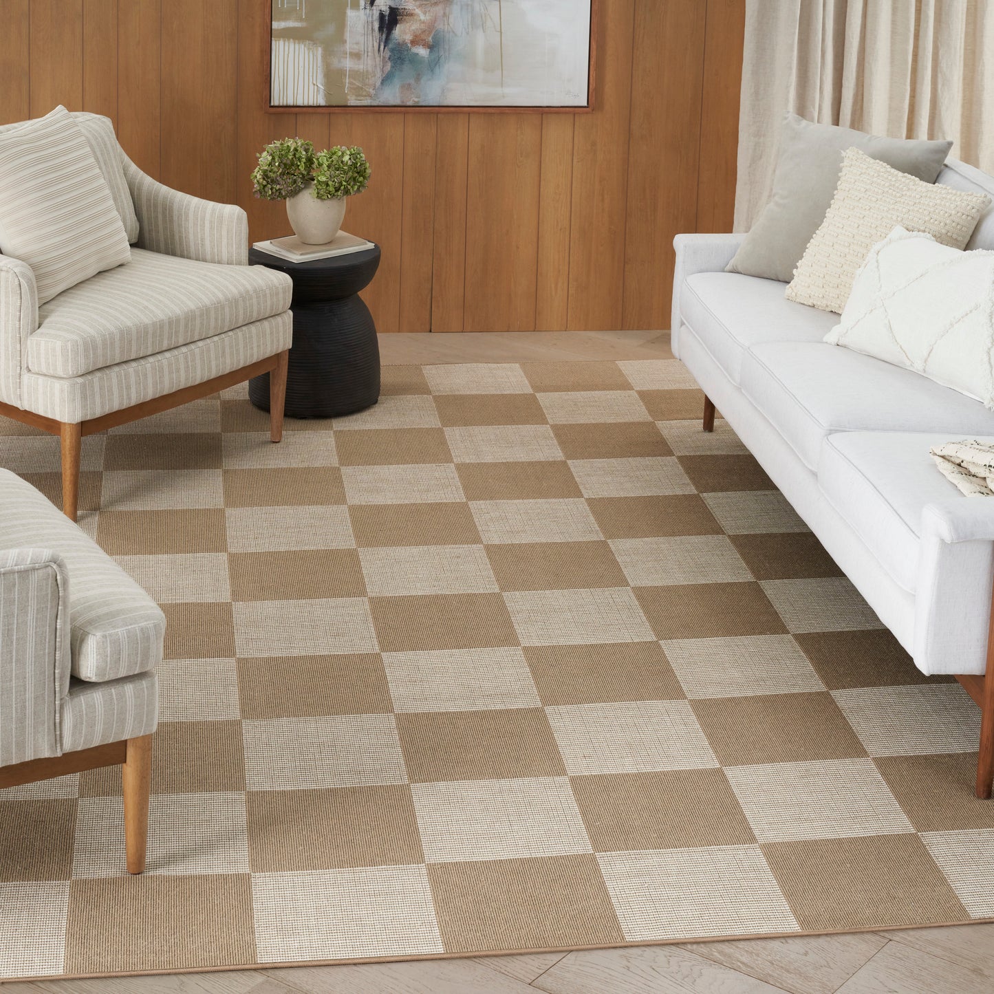 Living room with beige-brown checker rug, light striped armchairs, white sofa, wood paneled wall & potted plant.