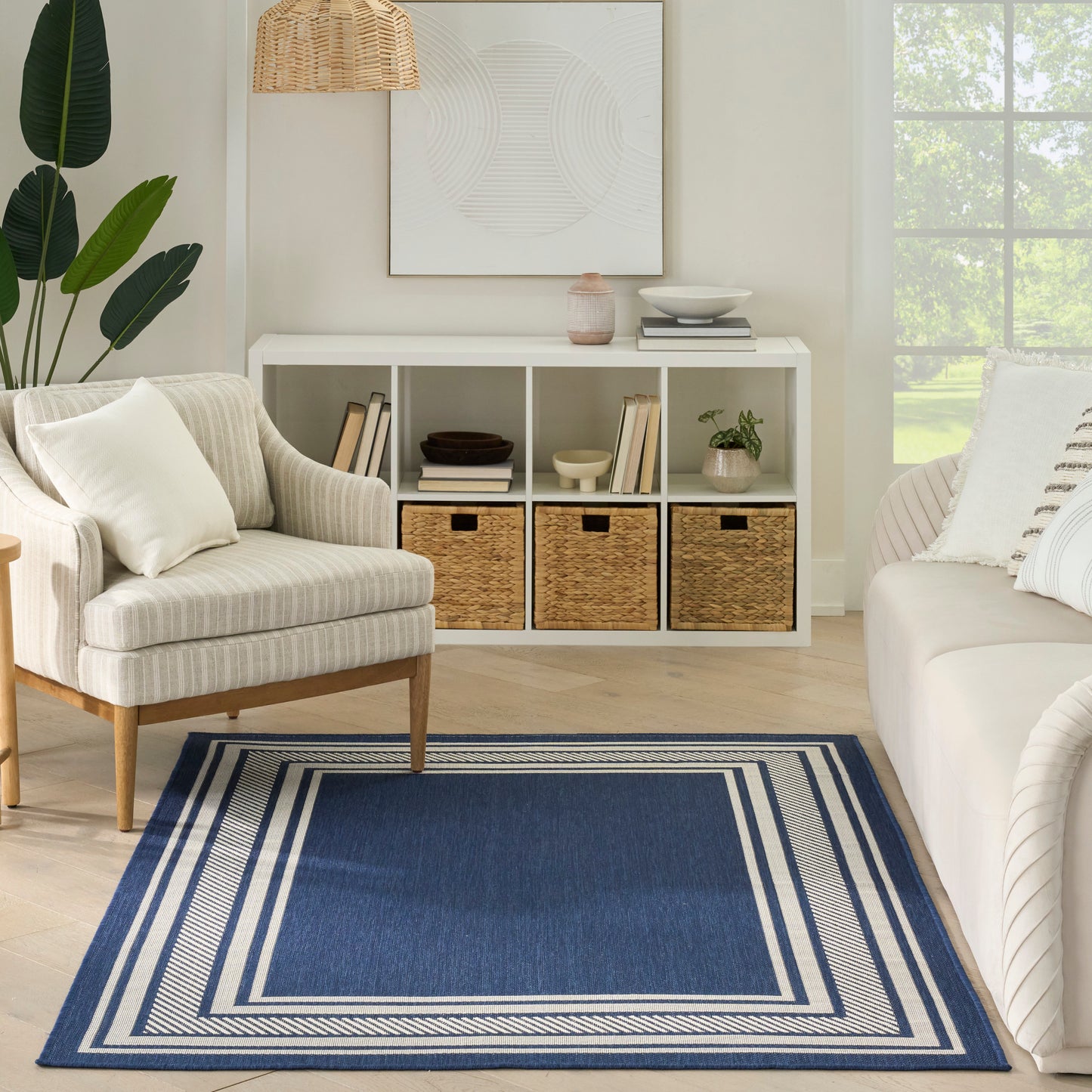 Coastal living room with white cubby storage, woven baskets, beige striped armchair, navy-blue bordered rug, greenery.