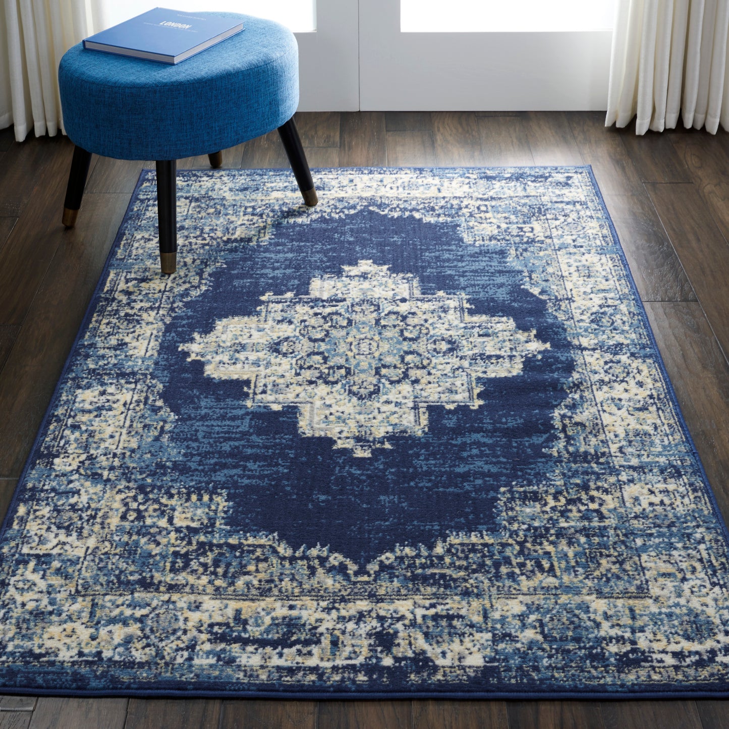 Navy blue area rug featuring a distressed ivory medallion center on dark wood floor, paired with a round blue ottoman.
