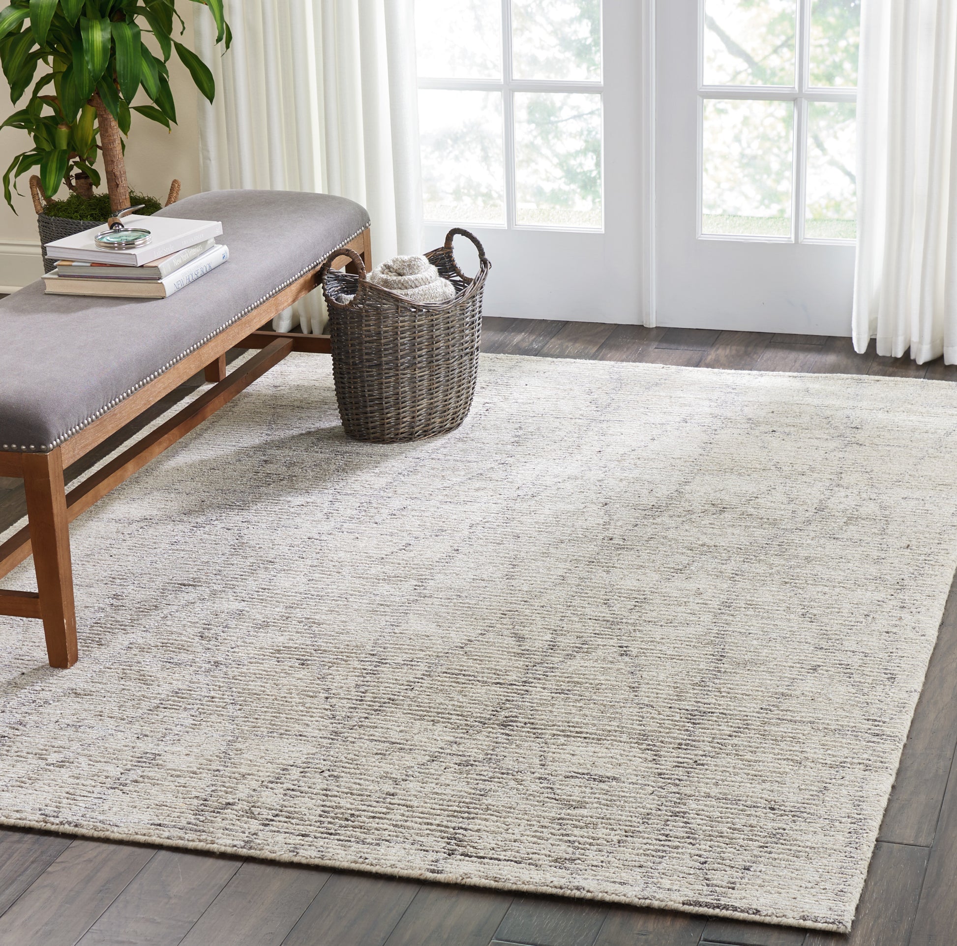 Beige textured area rug on dark wood floor with gray upholstered bench, wicker basket, potted plant, and large windows.